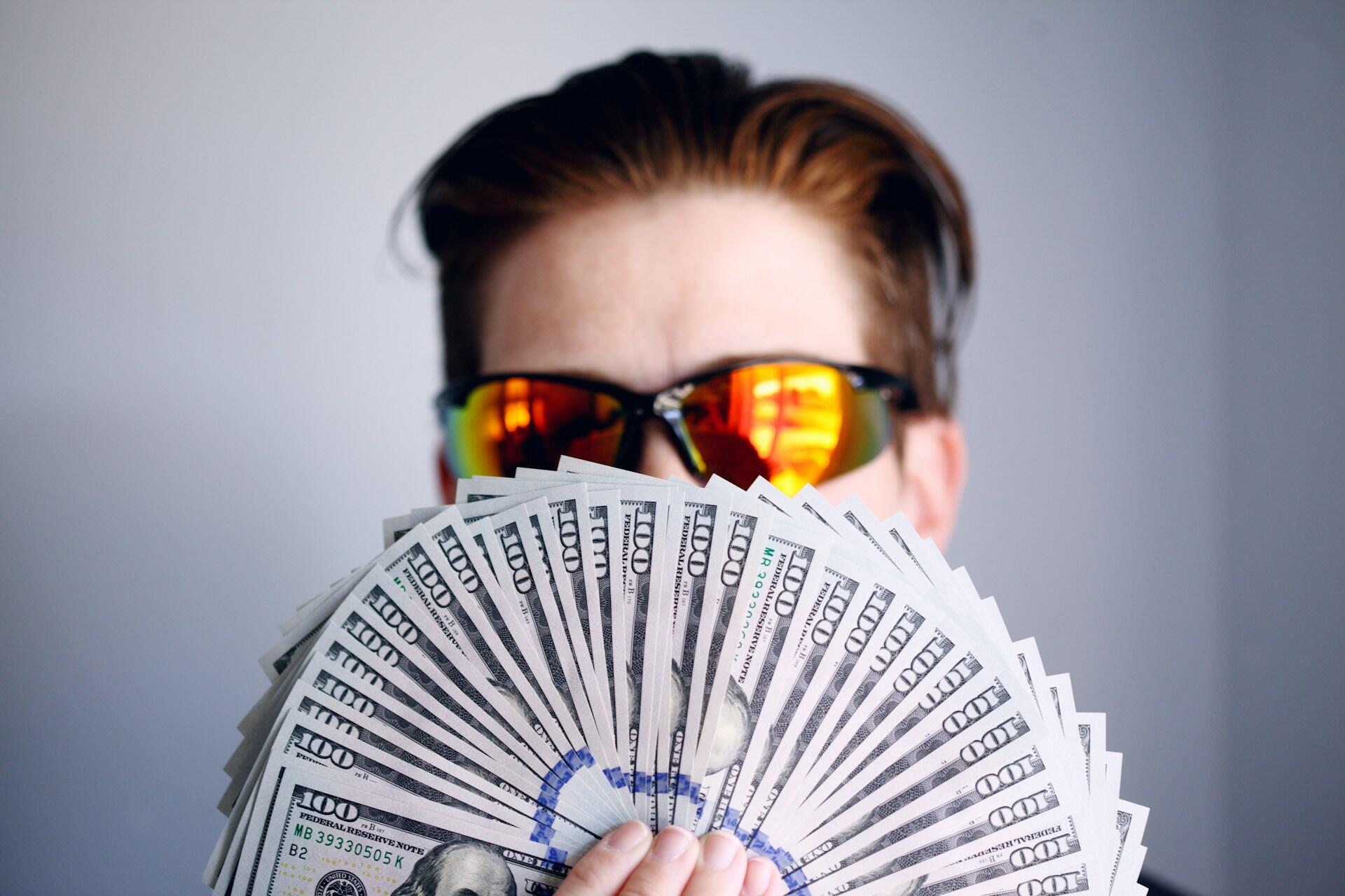 Człowiek w okularach przeciwsłonecznych z wachlarzem banknotów przed twarzą.