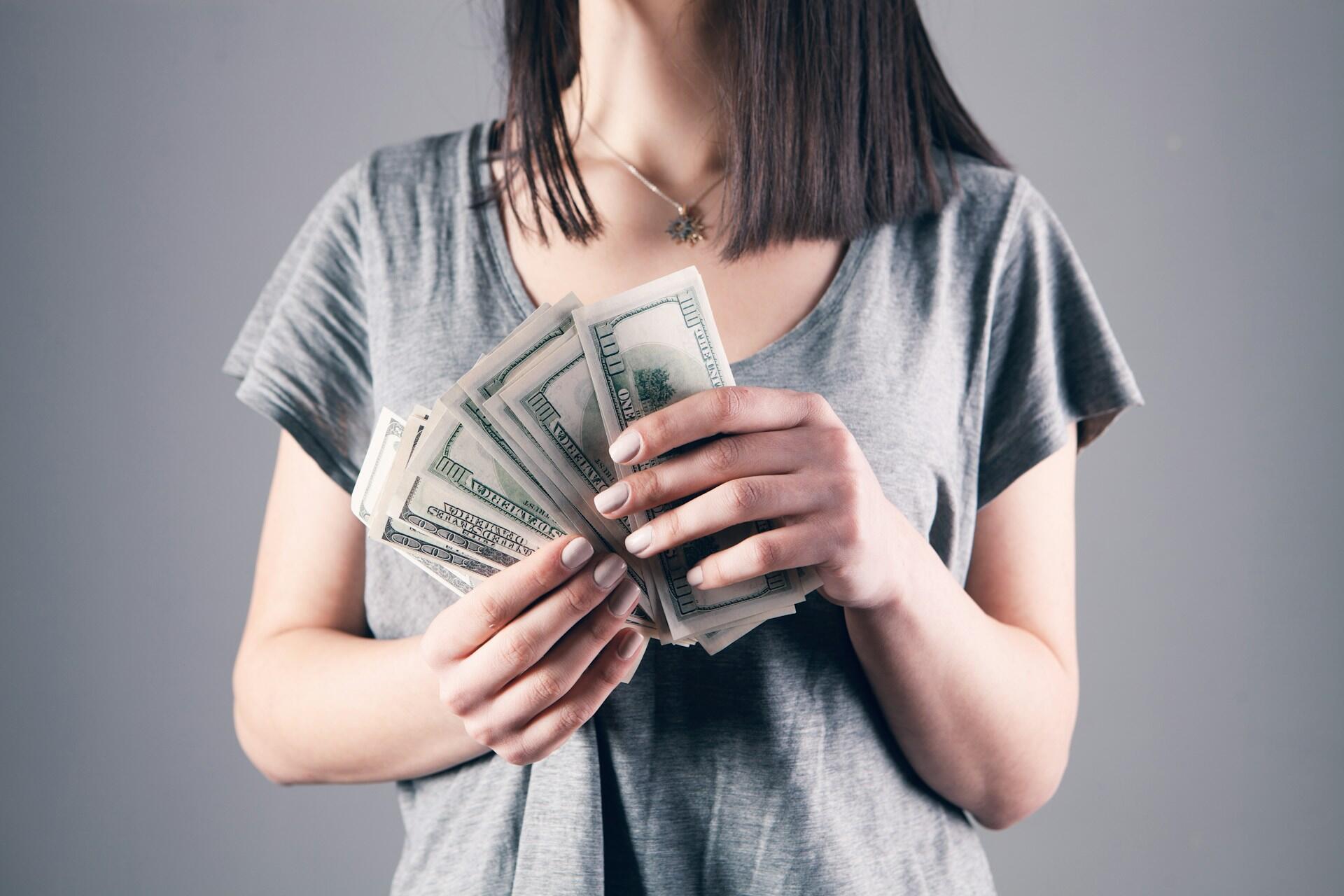 Kobieta w szarej koszulce z plikiem dolarów w rękach.