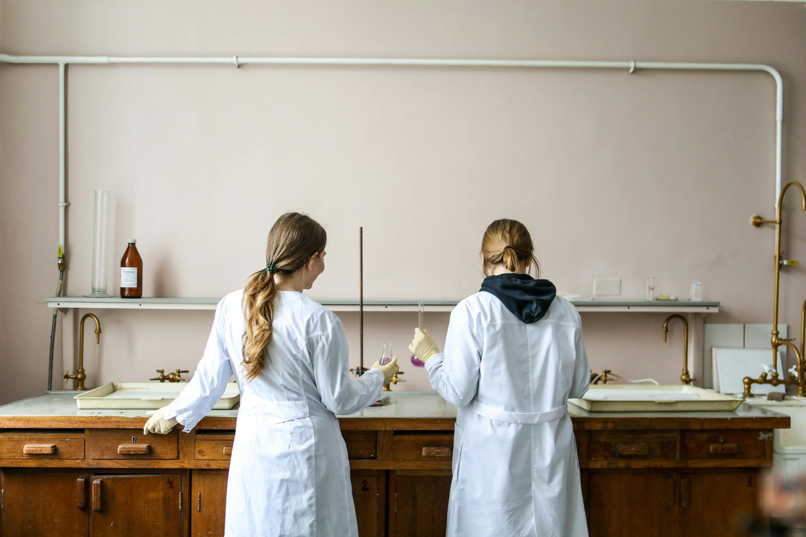 2 uczennice w fartuchach na lekcji chemii, stoją przy blacie z ze szkłem laboratoryjnym w dłoniach, fot. pexels.com