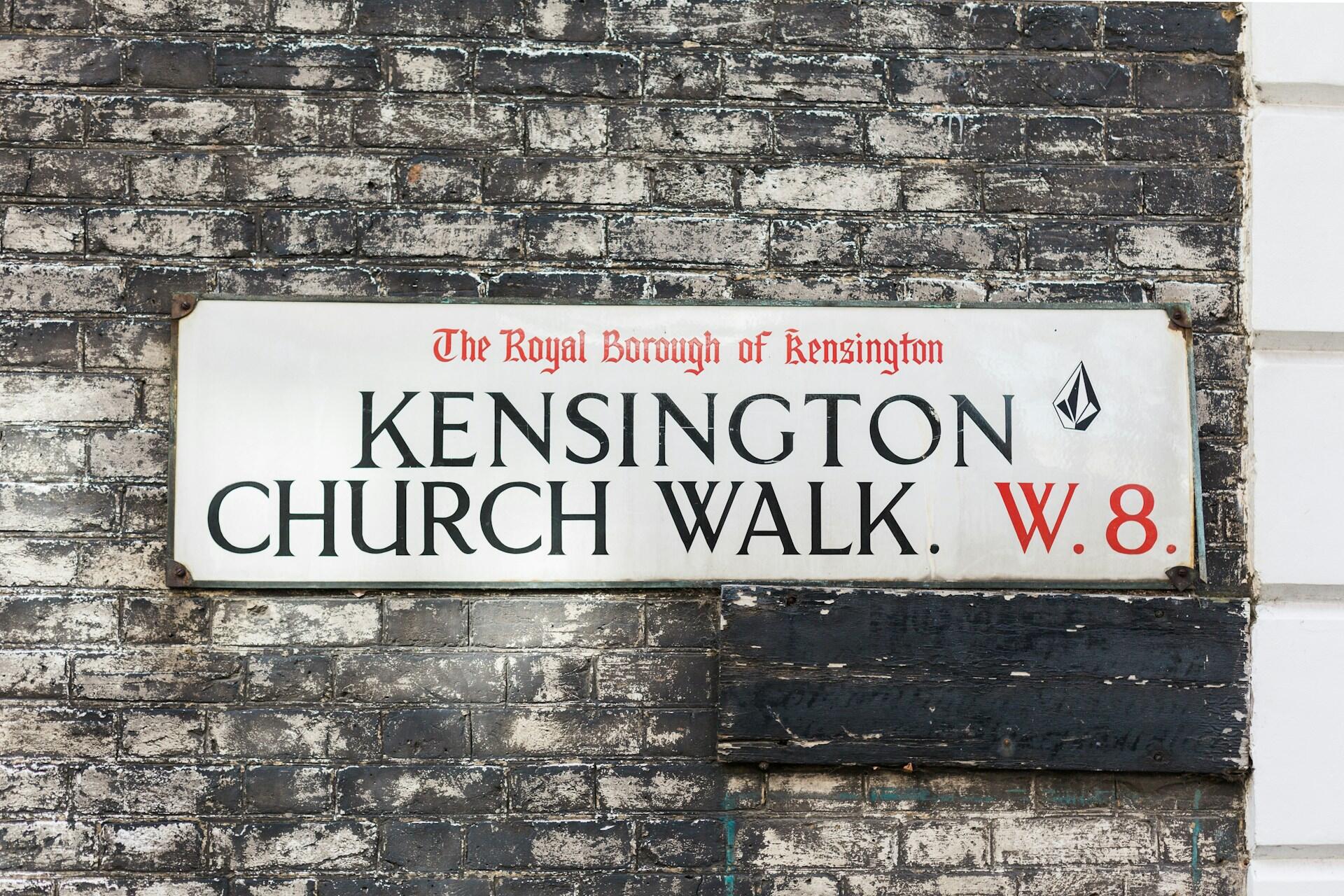 Zniszczony znak drogowy z napisem „Kensington Church Walk, W.8” otoczony teksturowaną ścianą z cegły w dzielnicy Kensington w Londynie.