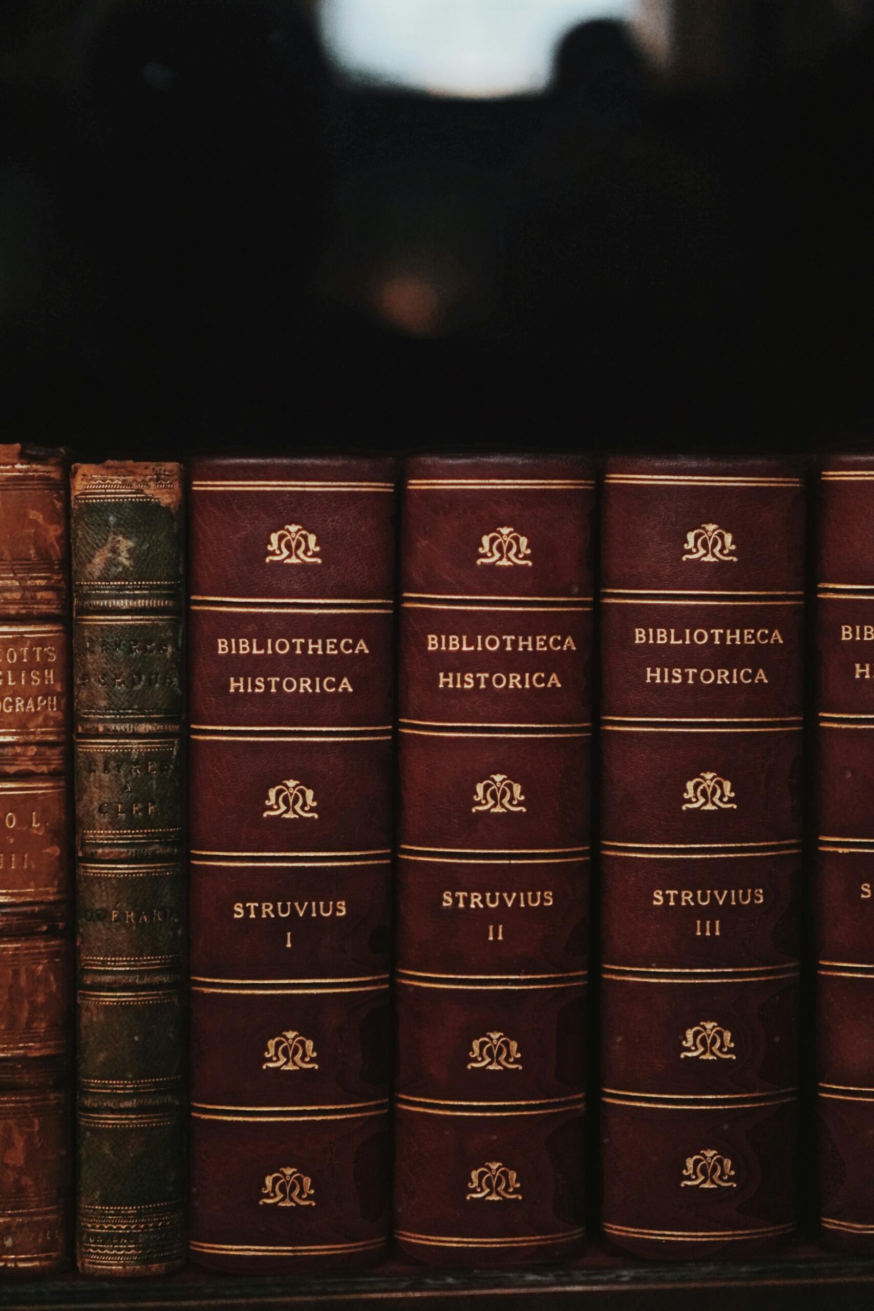 Fragment półki z zabytkowymi, elegancko oprawionymi książkami historycznymi w ciemnoczerwonych (bordo/brązowych) oprawach ze złoconymi tłoczeniami. Unsplash: Precious Madubuike