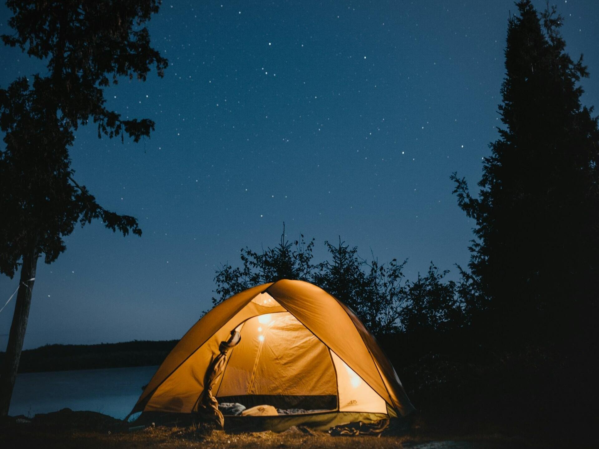Świecący na żółto namiot stoi nad spokojnym jeziorem pod rozgwieżdżonym niebem, otoczony drzewami, tworząc przytulną atmosferę kempingu.