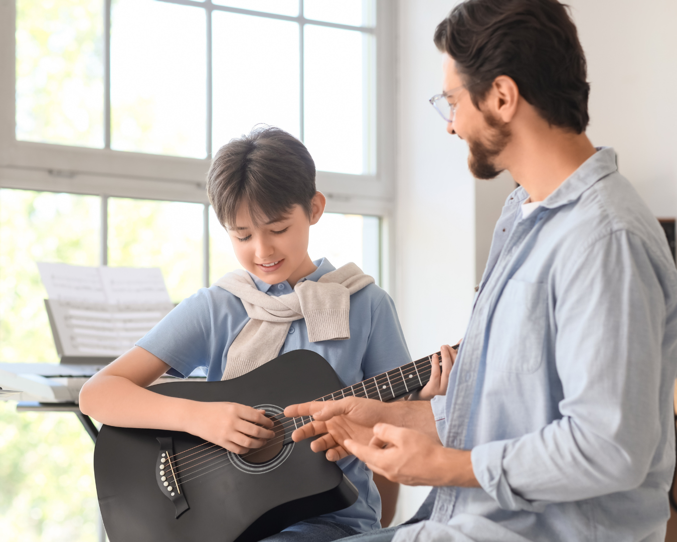 Nauczyciel pomaga młodemu uczniowi grać na czarnej gitarze podczas lekcji muzyki w jasnym pokoju z dużymi oknami.