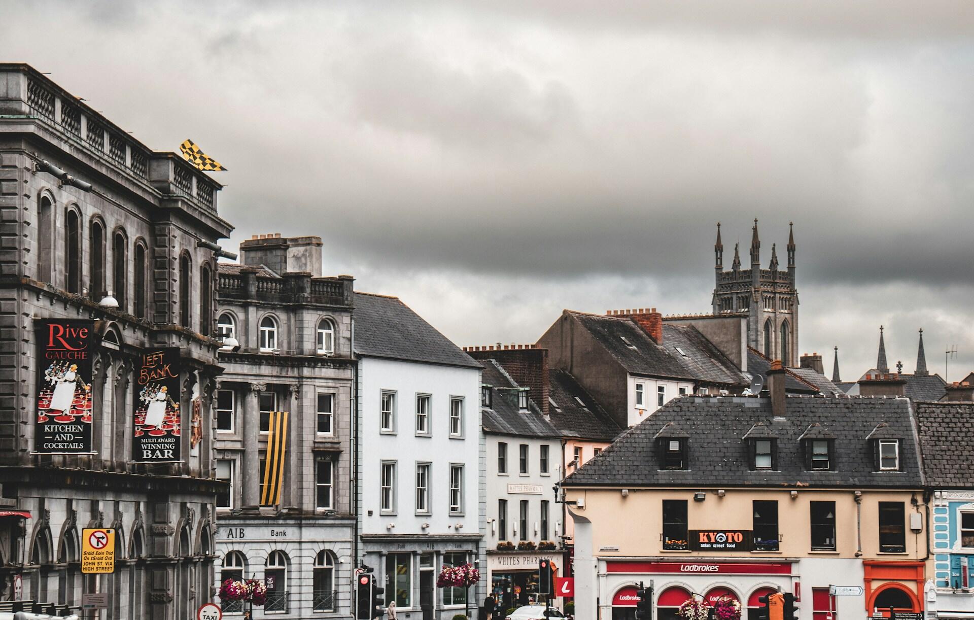 Widok na ryneczek z zabytkowymi budynkami, sklepami i wieżą kościoła pod zachmurzonym niebem.