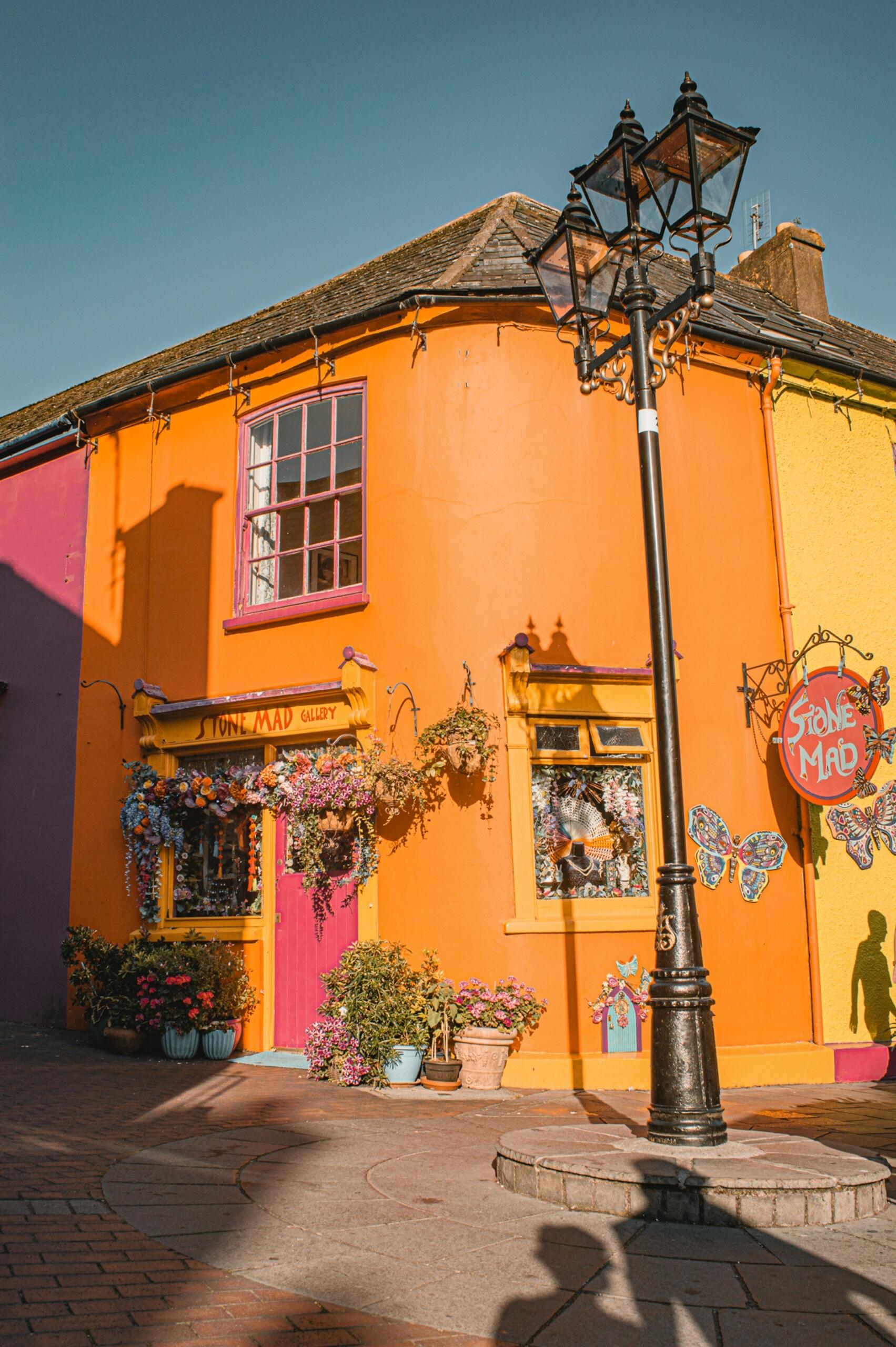 Jasny budynek z pomarańczowymi i żółtymi ścianami, ozdobiony kwiatami. W pobliżu stoi latarnia, rzucająca cień na chodnik.