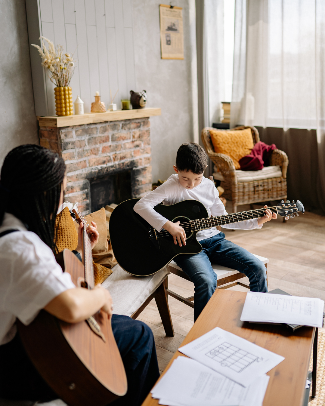 Osoba uczy dziecko gry na gitarze w przytulnym, dobrze oświetlonym salonie z rustykalnym kominkiem i rozłożonymi nutami