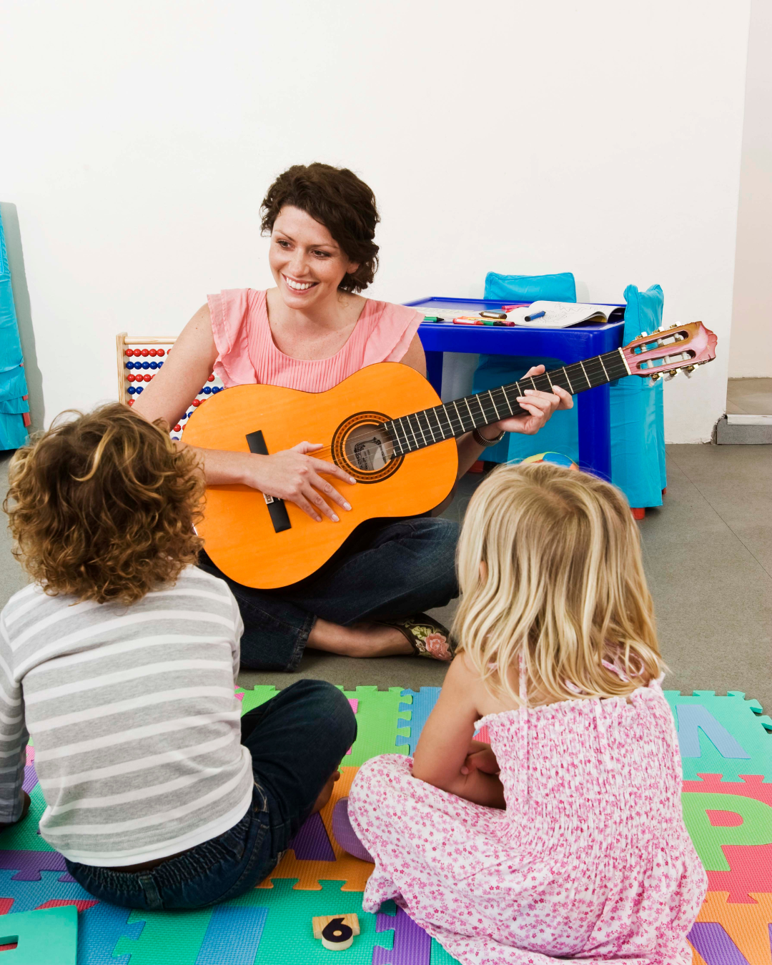 Kobieta gra na gitarze, siedząc na kolorowej macie piankowej, angażując dwoje dzieci, które siedzą przed nią z uwagą