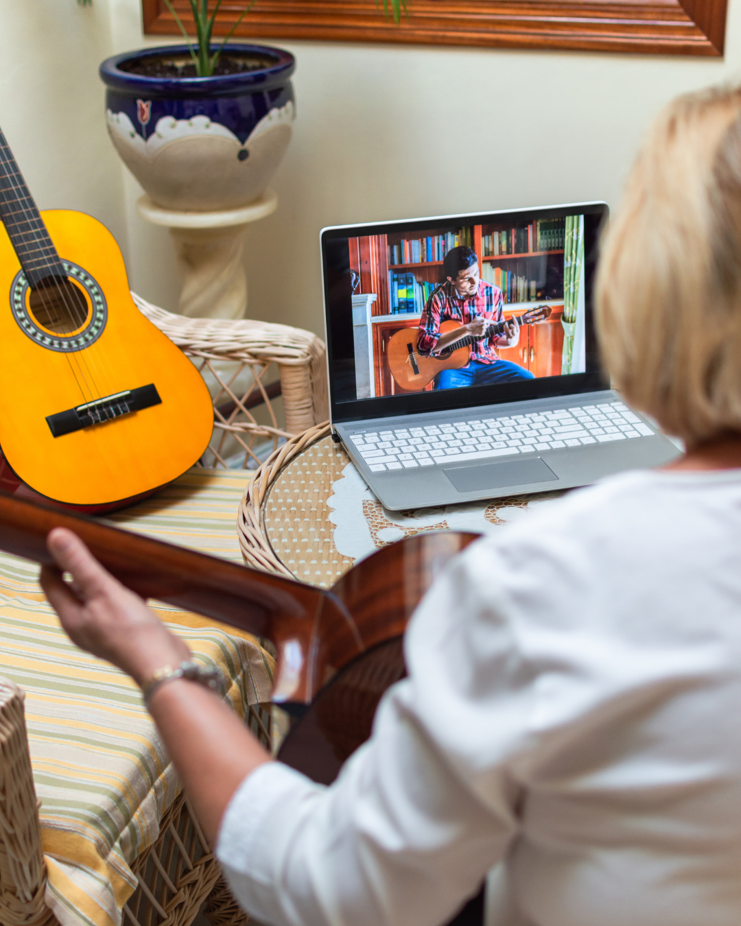 Osoba uczy się gry na gitarze online, siedząc przy stole z laptopem wyświetlającym lekcję, obok której znajduje się kolejna gitara.