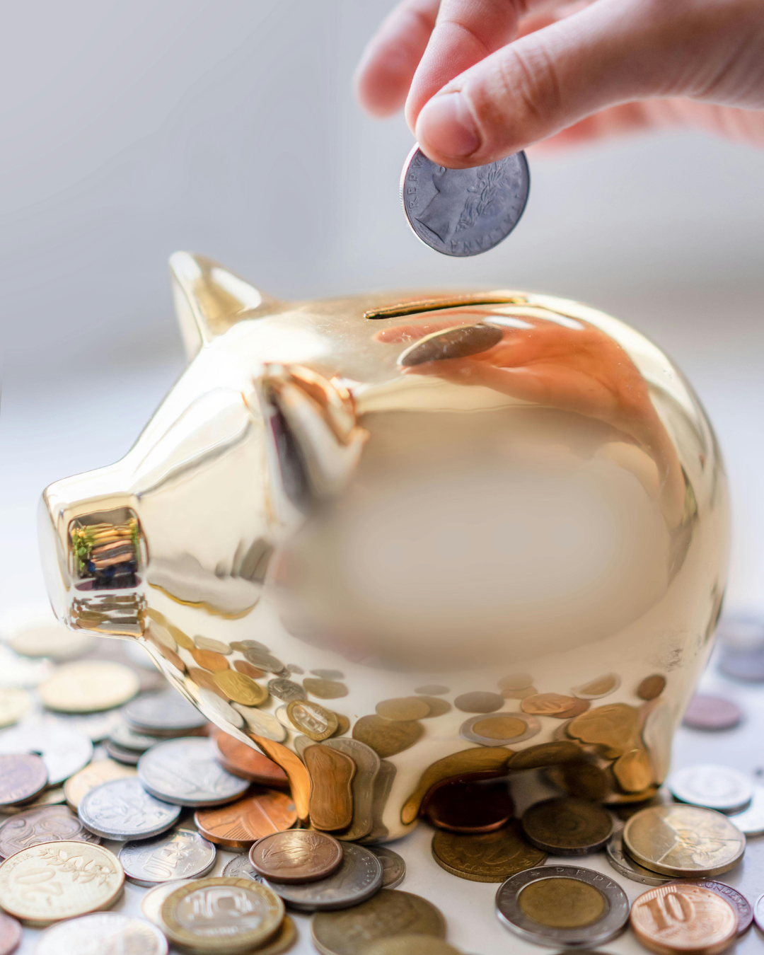 Ręka wrzuca monetę do błyszczącej złotej skarbonki otoczonej różnymi monetami na jasnym podłożu.