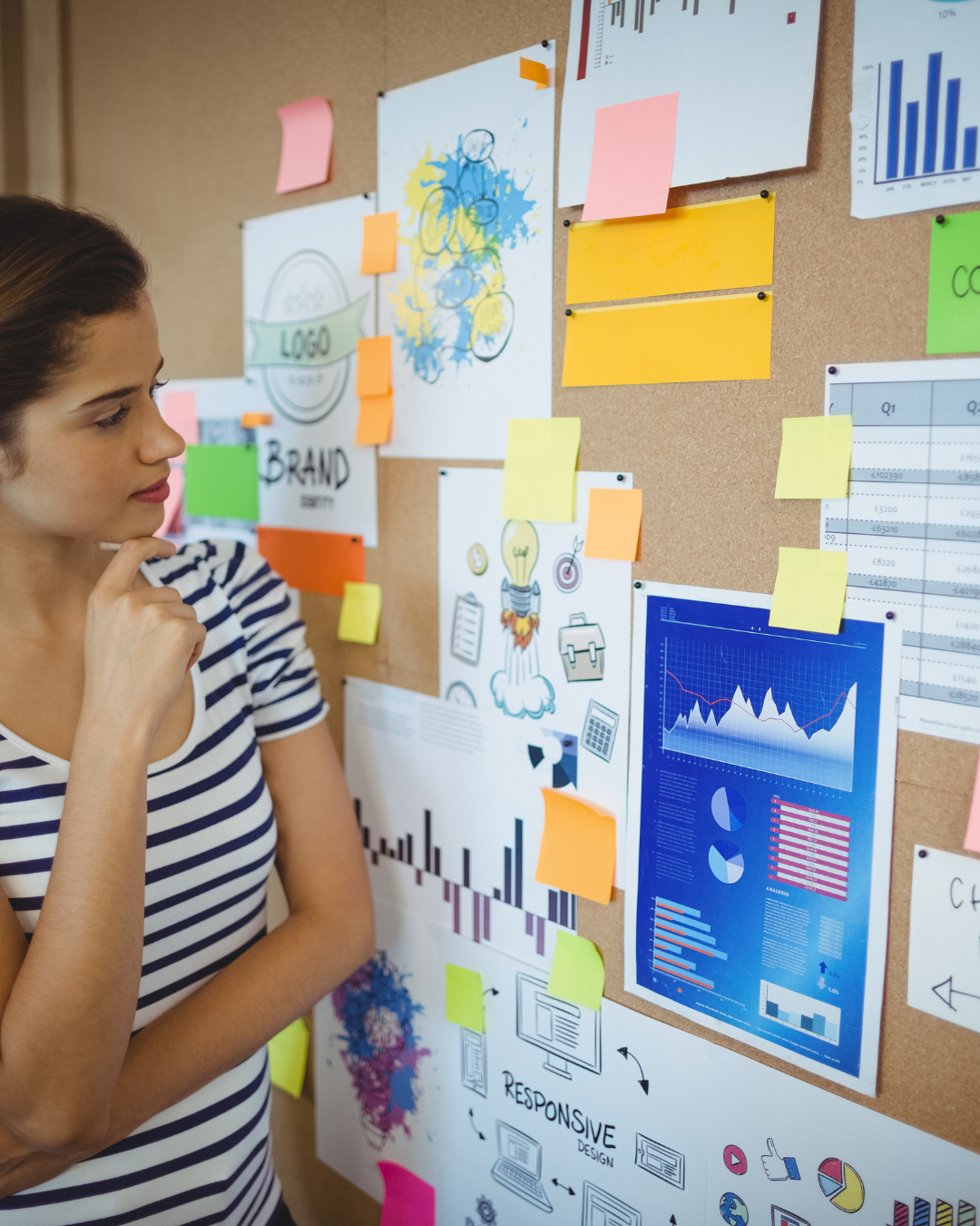 Osoba stojąca przy tablicy korkowej pokrytej kolorowymi karteczkami samoprzylepnymi, wykresami i szkicami projektowymi, pogrążona w głębokiej zadumie.