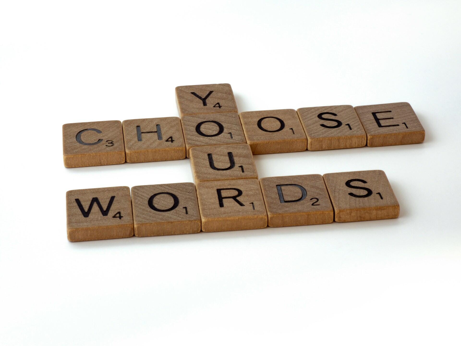 Płytki do gry Scrabble układają się w napis „Choose Your Words”, ułożony w kształt krzyża na białym tle.
