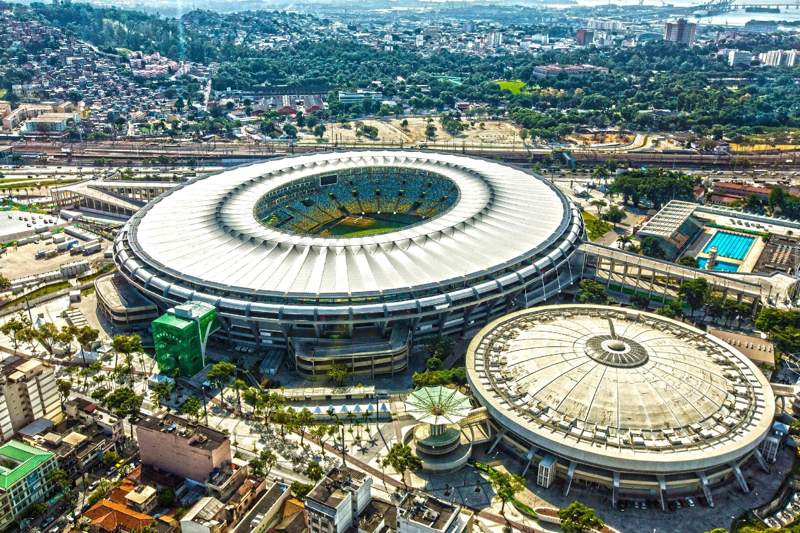 Widok z lotu ptaka na stadion Maracanã i sąsiednią halę sportową w Rio de Janeiro, otoczone zabudową miejską i terenami zielonymi.