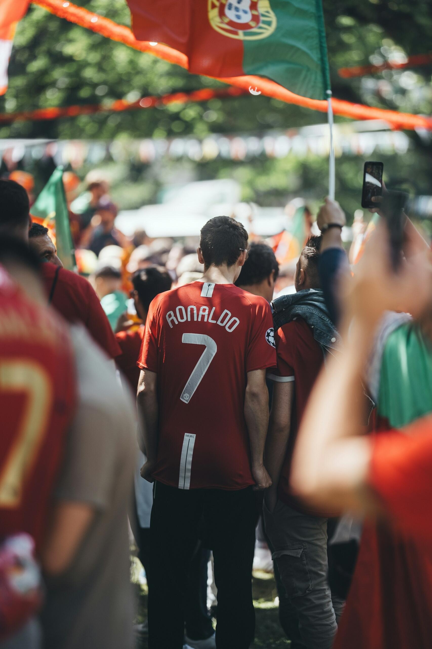 Tłum kibiców piłkarskich na świeżym powietrzu; jeden z nich ma na sobie czerwoną koszulkę Ronaldo z numerem 7, a nad nimi powiewa portugalska flaga.