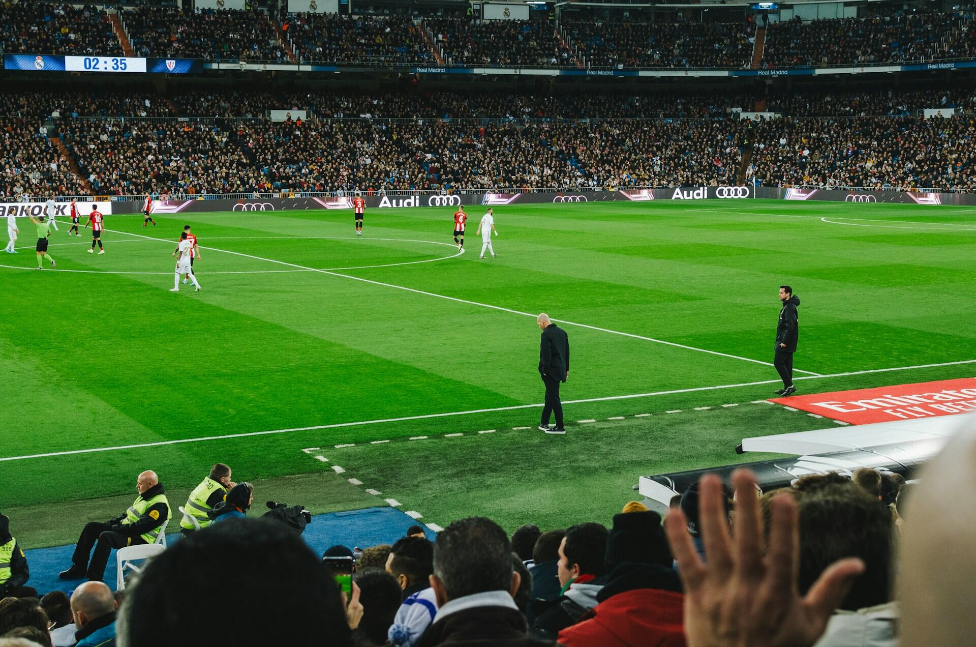 Mecz piłki nożnej na stadionie, z piłkarzami na boisku, trenerami przy linii bocznej i tłumem kibiców na trybunach.