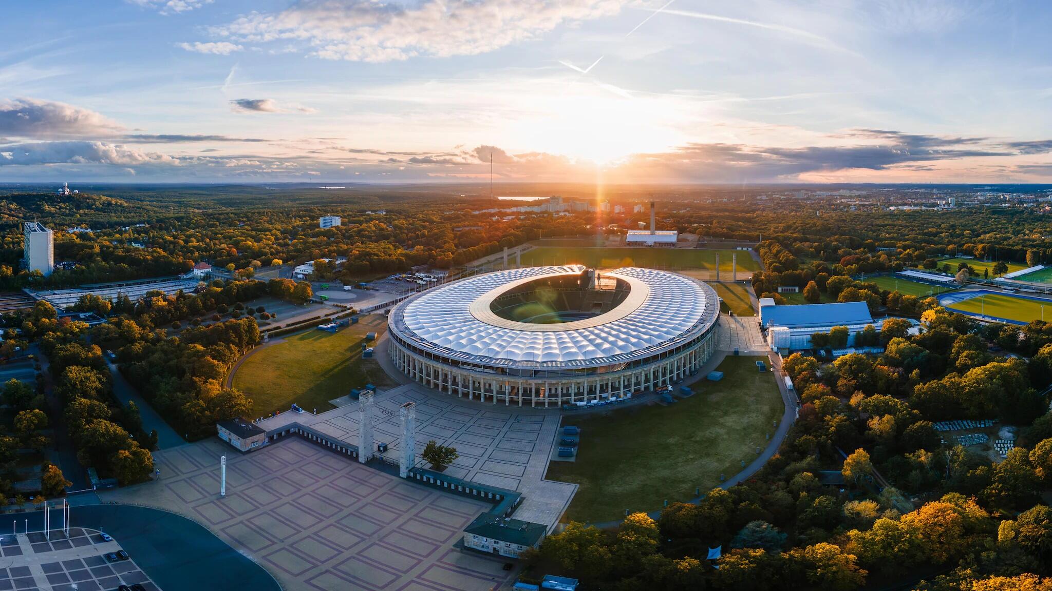 Widok z lotu ptaka na duży, okrągły stadion otoczony drzewami i otwartymi polami o zachodzie słońca przy bezchmurnym niebie.