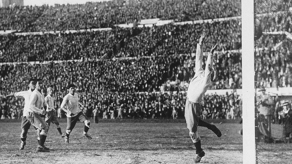 Czarno-białe zdjęcie bramkarza piłkarskiego wyciągającego rękę, by zablokować piłkę, podczas gdy gracze i zgromadzeni na stadionie kibice obserwują tę akcję.