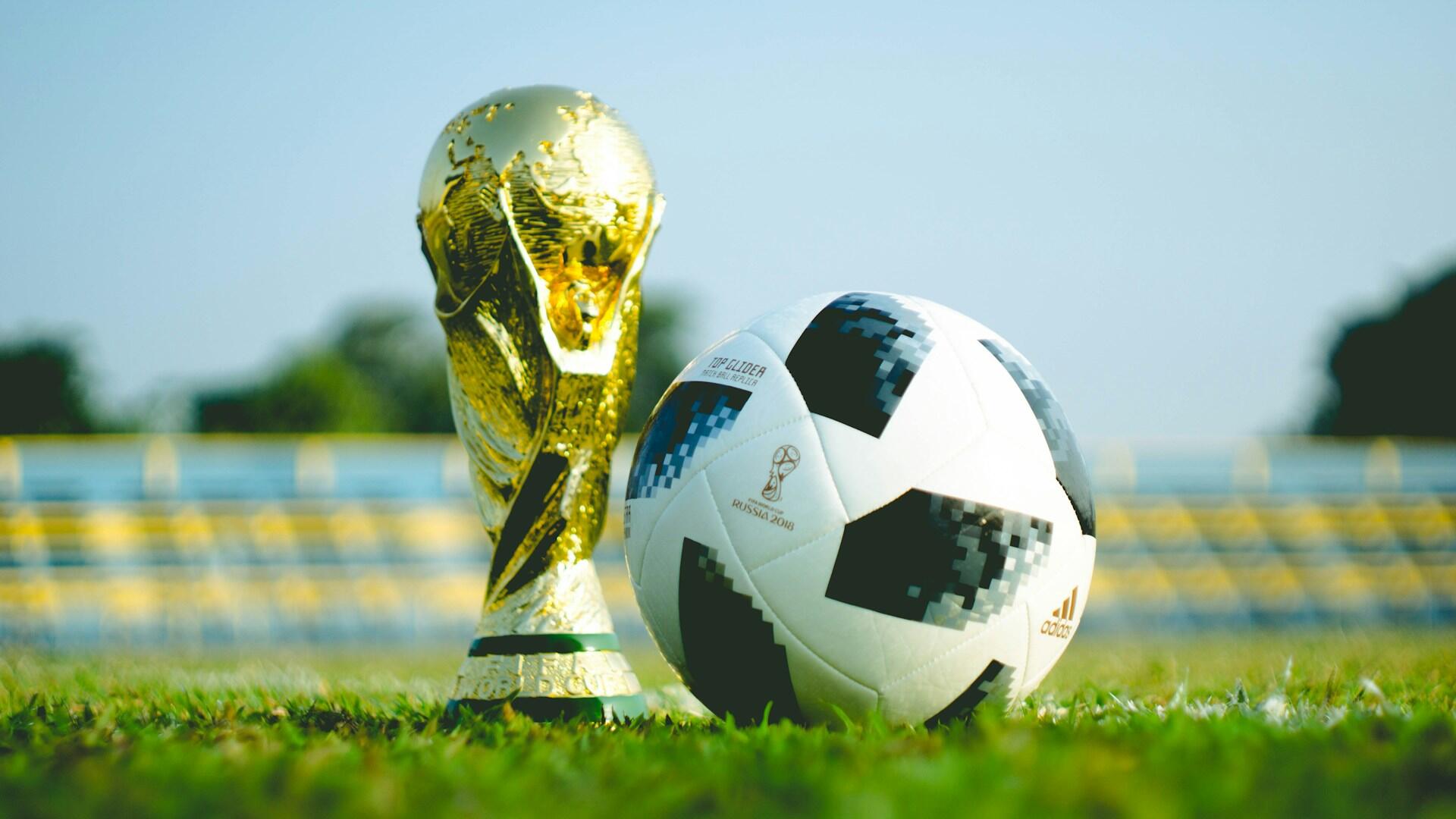 Trofeum mistrzostw świata i oficjalna piłka z mistrzostw świata w Rosji w 2018 roku na trawiastym boisku, a w tle zamazane trybuny stadionu.