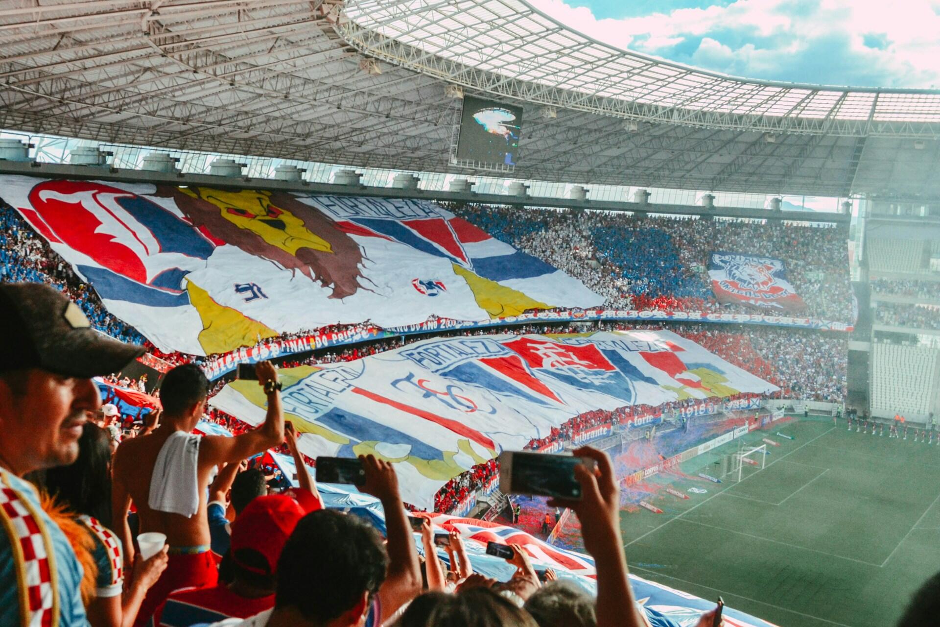 Tłum kibiców na stadionie trzymających duże, kolorowe transparenty i robiących zdjęcia podczas meczu piłki nożnej.