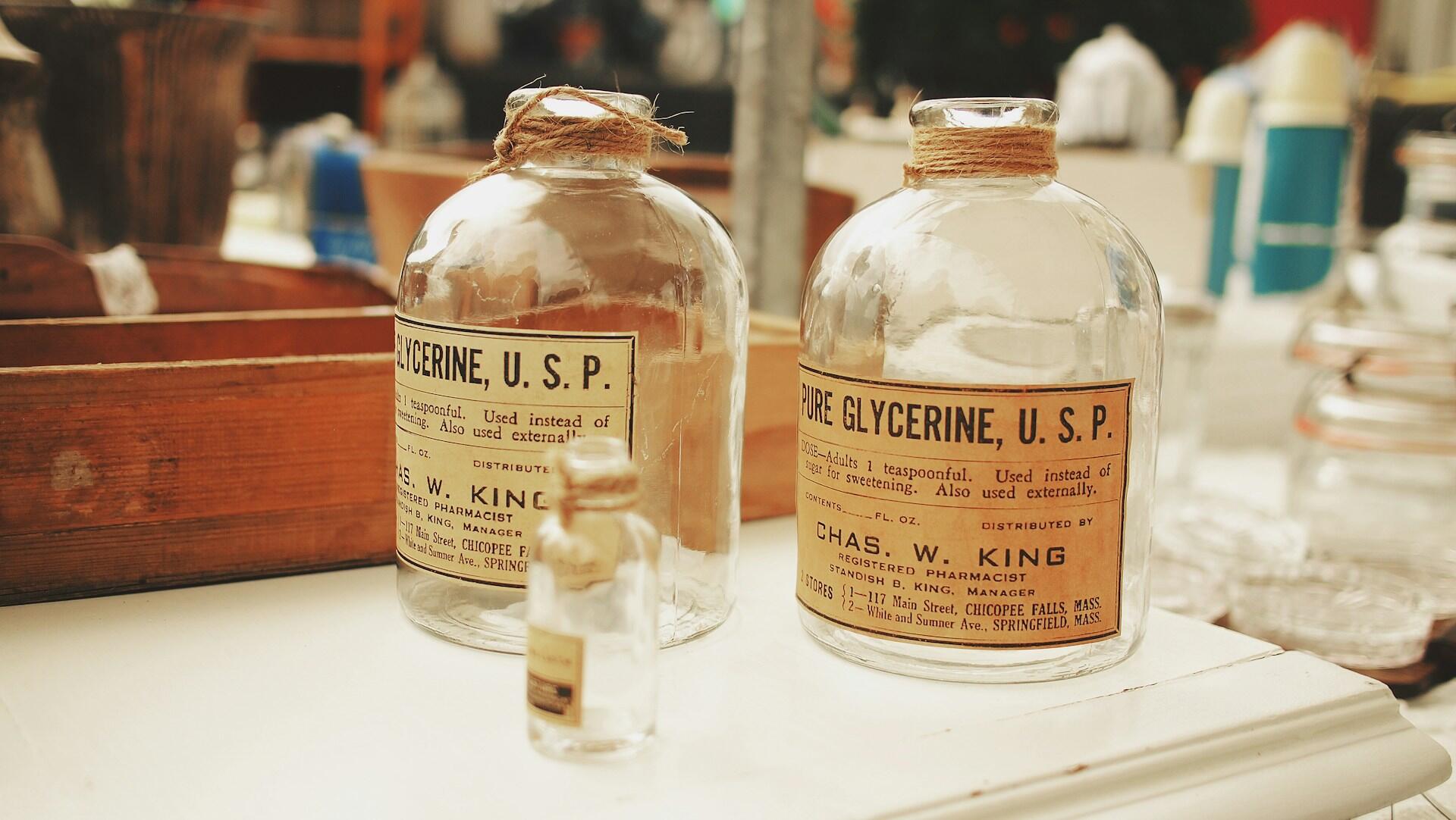 Dwie zabytkowe szklane butelki z napisem „Pure Glycerine, U.S.P.” na białym tle, a w tle drewniane pudełko.