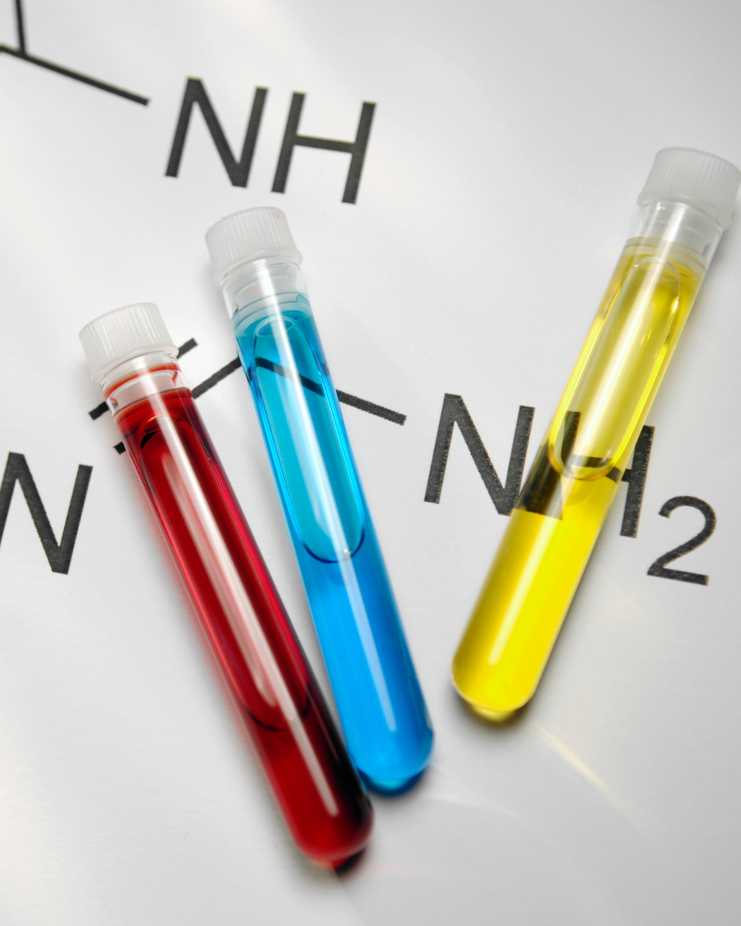 Trzy probówki wypełnione czerwonym, niebieskim i żółtym płynem na kartce z wzorem chemicznym zawierającym grupy NH i NH₂.