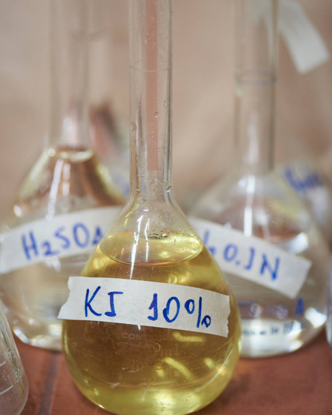 Zbliżenie na opatrzone etykietami szklane kolby zawierające roztwory chemiczne, ze szczególnym uwzględnieniem kolby z napisem „KI 10%”, w której znajduje się żółty płyn.