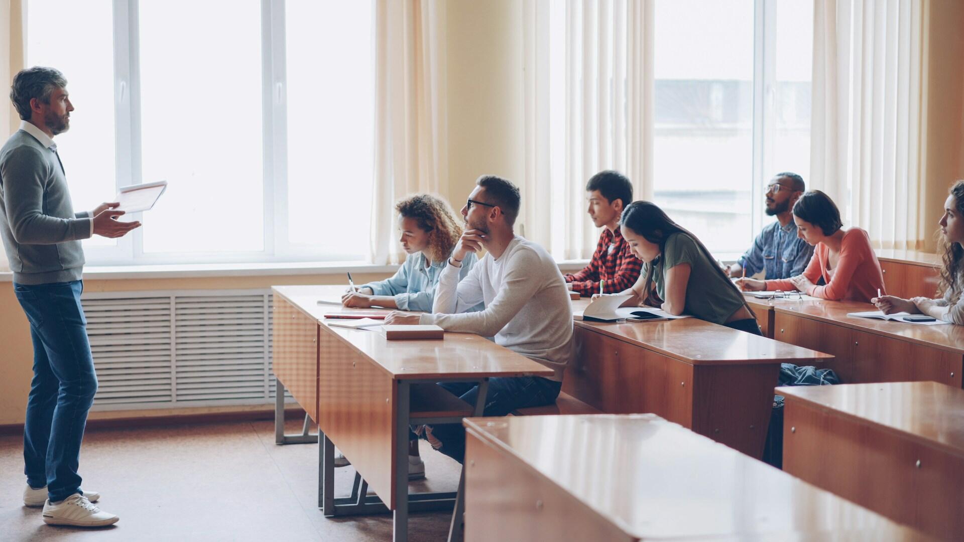 Profesor w swobodnym stroju rozmawia z grupą studentów siedzących przy stołach w sali wykładowej i robiących notatki. Widoczna jest duża sala wykładowa z biurkami, krzesłami i dużymi oknami.