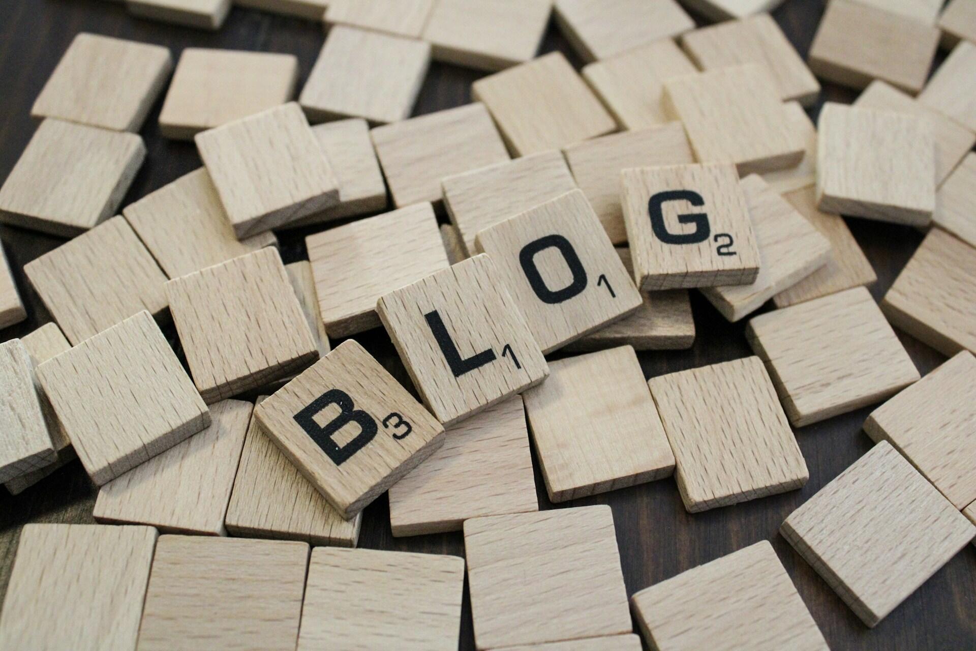 Drewniane płytki z literami i punktami tworzące napis "Blog" jak do gry w Scrabble leżące na odwróconych płytkach. Unsplash: Nadiia Ganzhyi