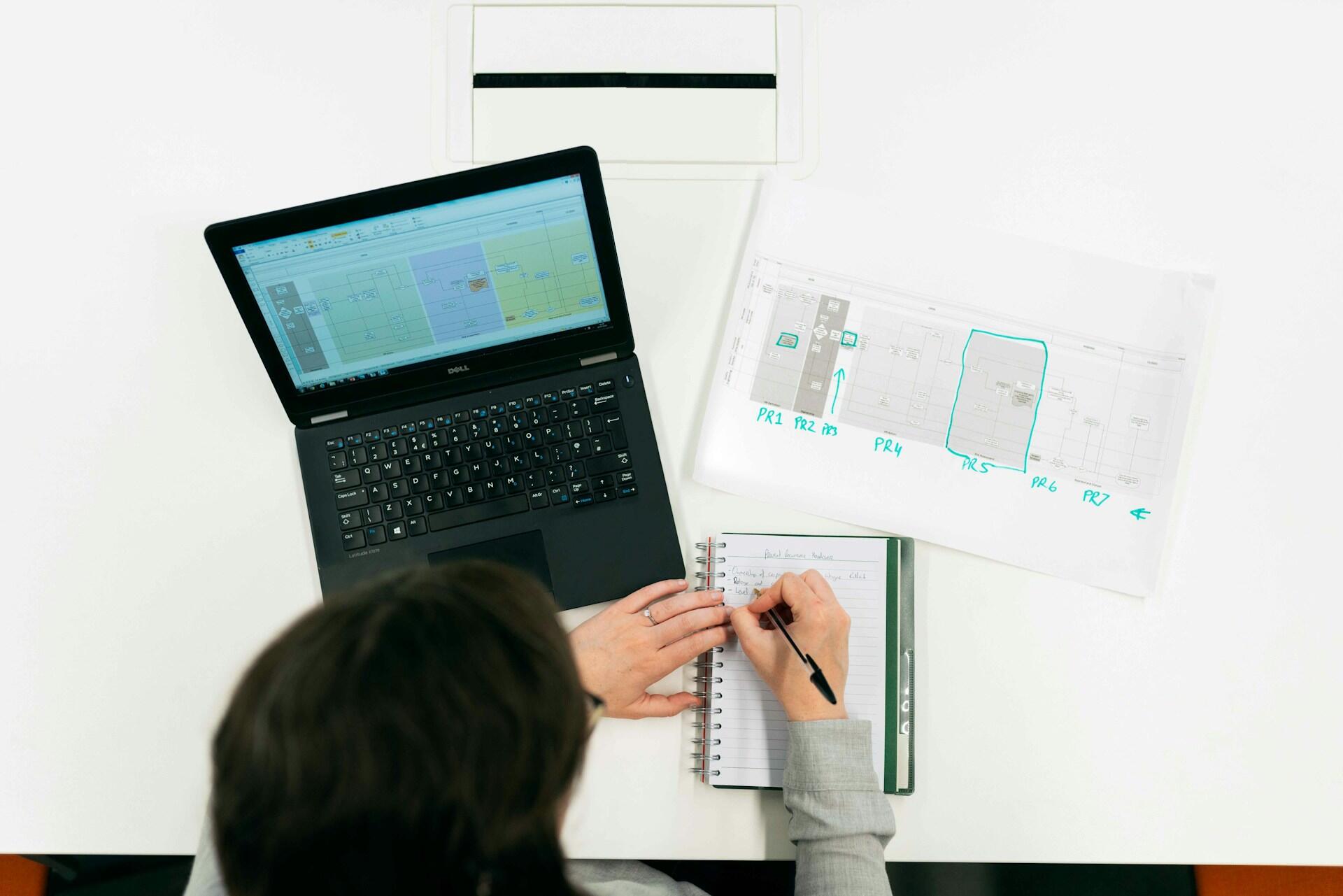 Kobieta inżynier elektryk siedzi przy biurku, na którym stoi laptop; pisze długopisem w notesie, obok ma wydruk z danymi. Unsplash: ThisisEngineering