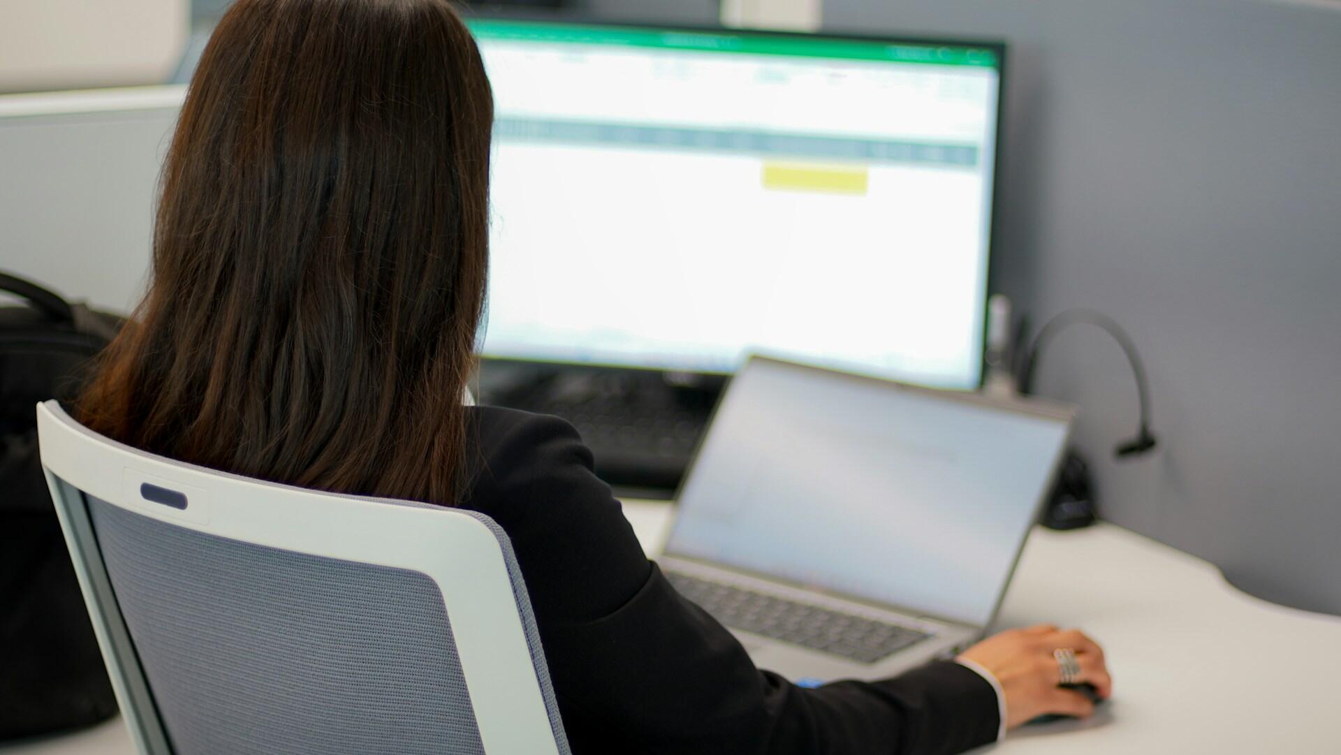 Kobieta siedząca tyłem do obiektywu, wpatrująca się w ekran komputera z tabelami Excel.