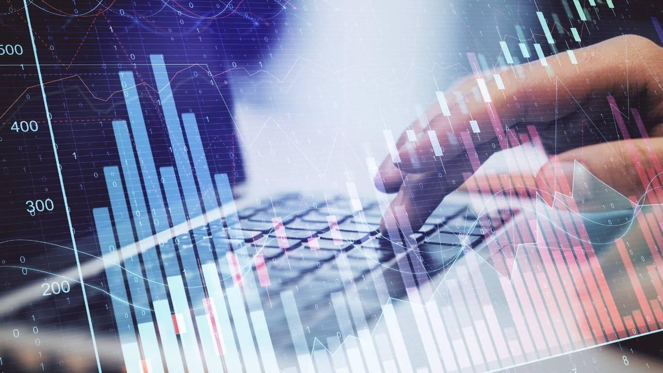 Ręce piszące na klawiaturze laptopa, na tle których widoczne są cyfrowe wykresy finansowe i diagramy danych przedstawiające analizę rynkową.