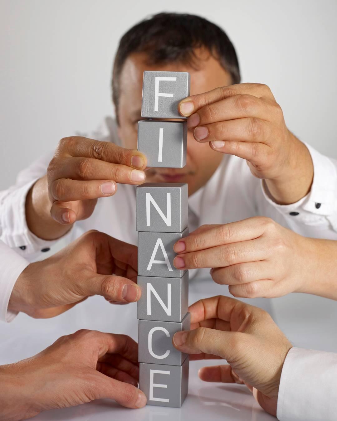 Wiele rąk współpracuje, układając klocki tworzące słowo „FINANCE” przed mężczyzną.