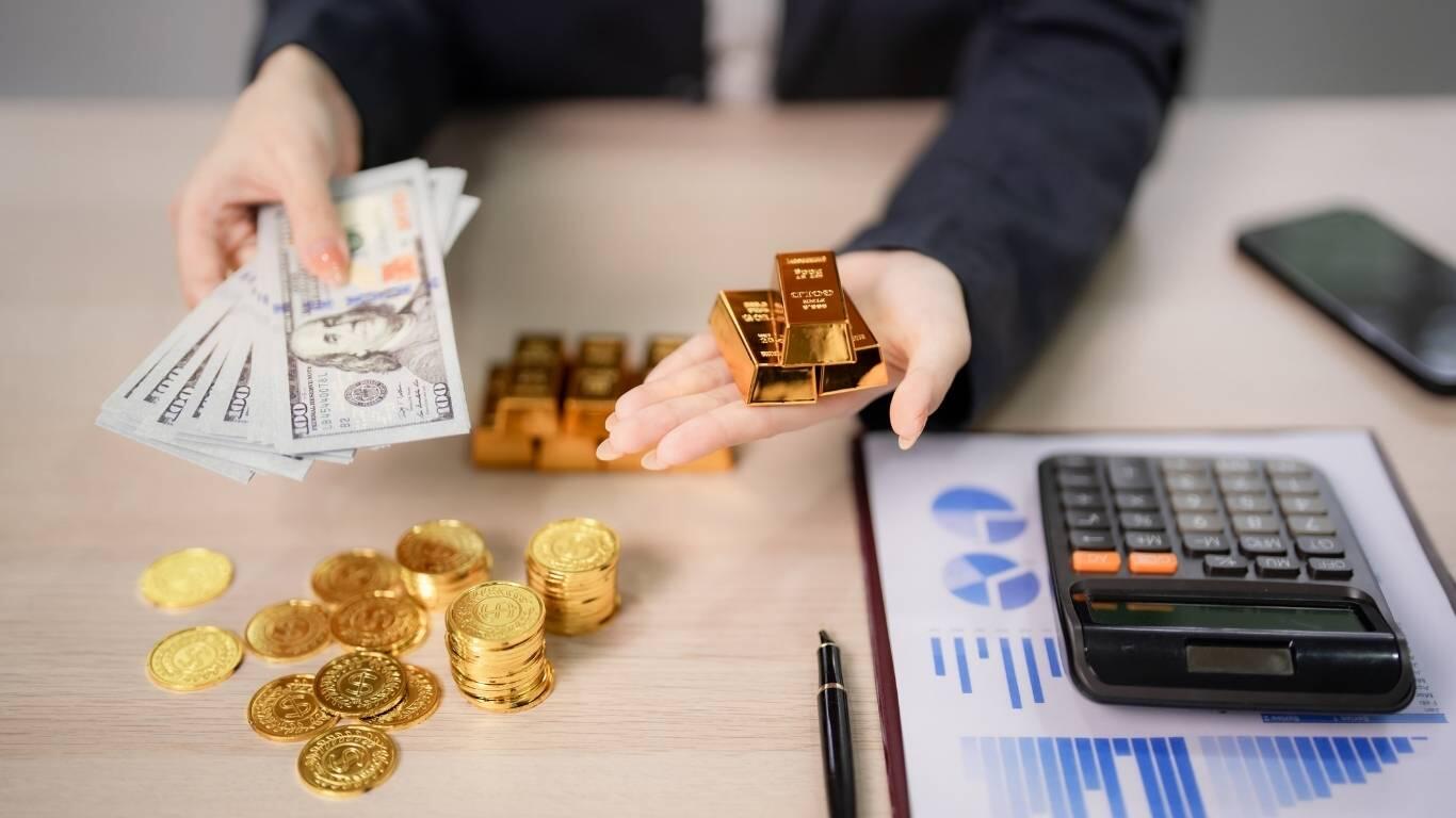 Osoba trzymająca w jednej ręce sztabki złota, a w drugiej banknoty dolarowe; na stole leżą złote monety, kalkulator i wykresy finansowe.