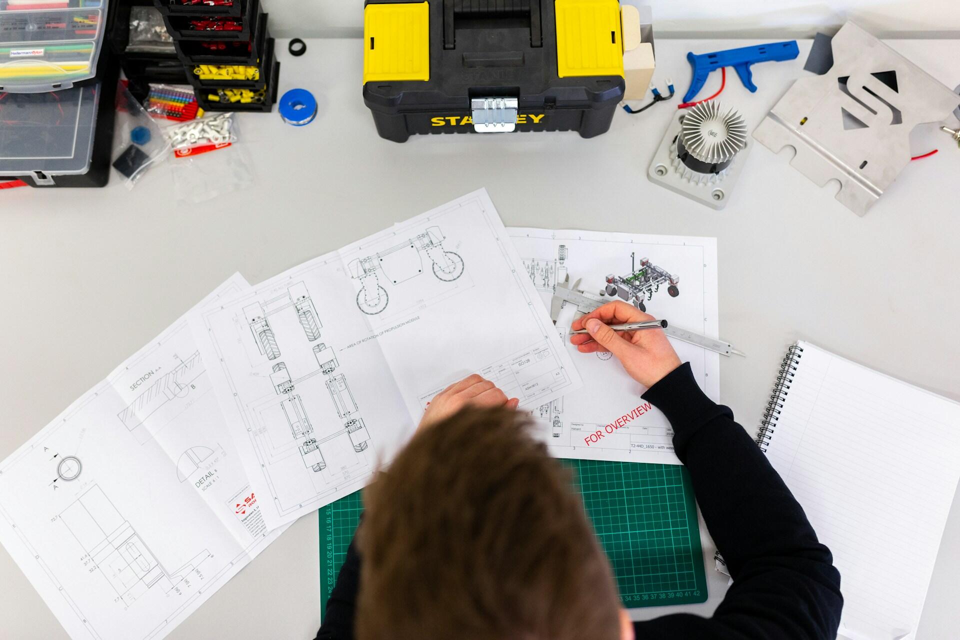 Mężczyzna pracuje nad projekt urządzeń, trzyma w ręku długopis, a na stole leżą narzędzia. Unsplash: ThisisEngineering