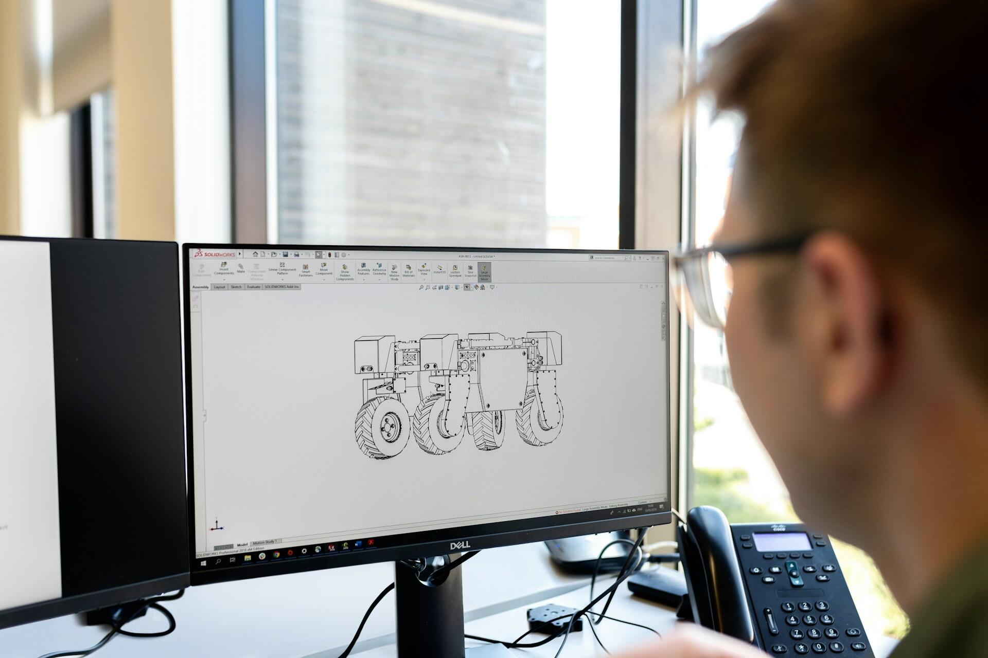 Inżynier mechanik siedzi przed ekranem komputera projektując maszynę, na biurku stoi telefon