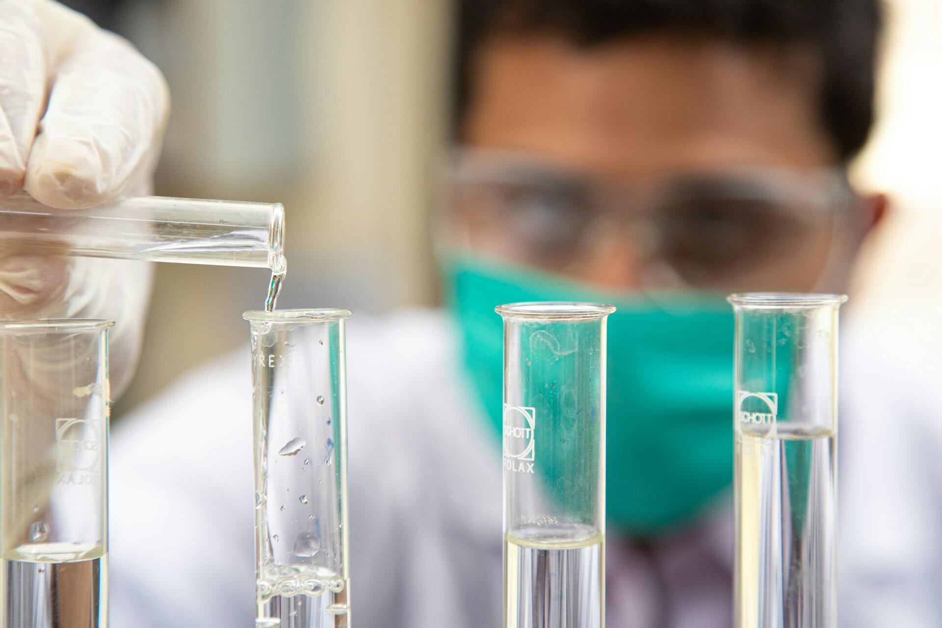 Pracownik laboratoryjny wykonuje doświadczenia chemicznego - ręka w białej rękawiczce ochronnej przelewa przezroczystą ciecz z jednej probówki do drugiej. Unsplash: Talha Hassan
