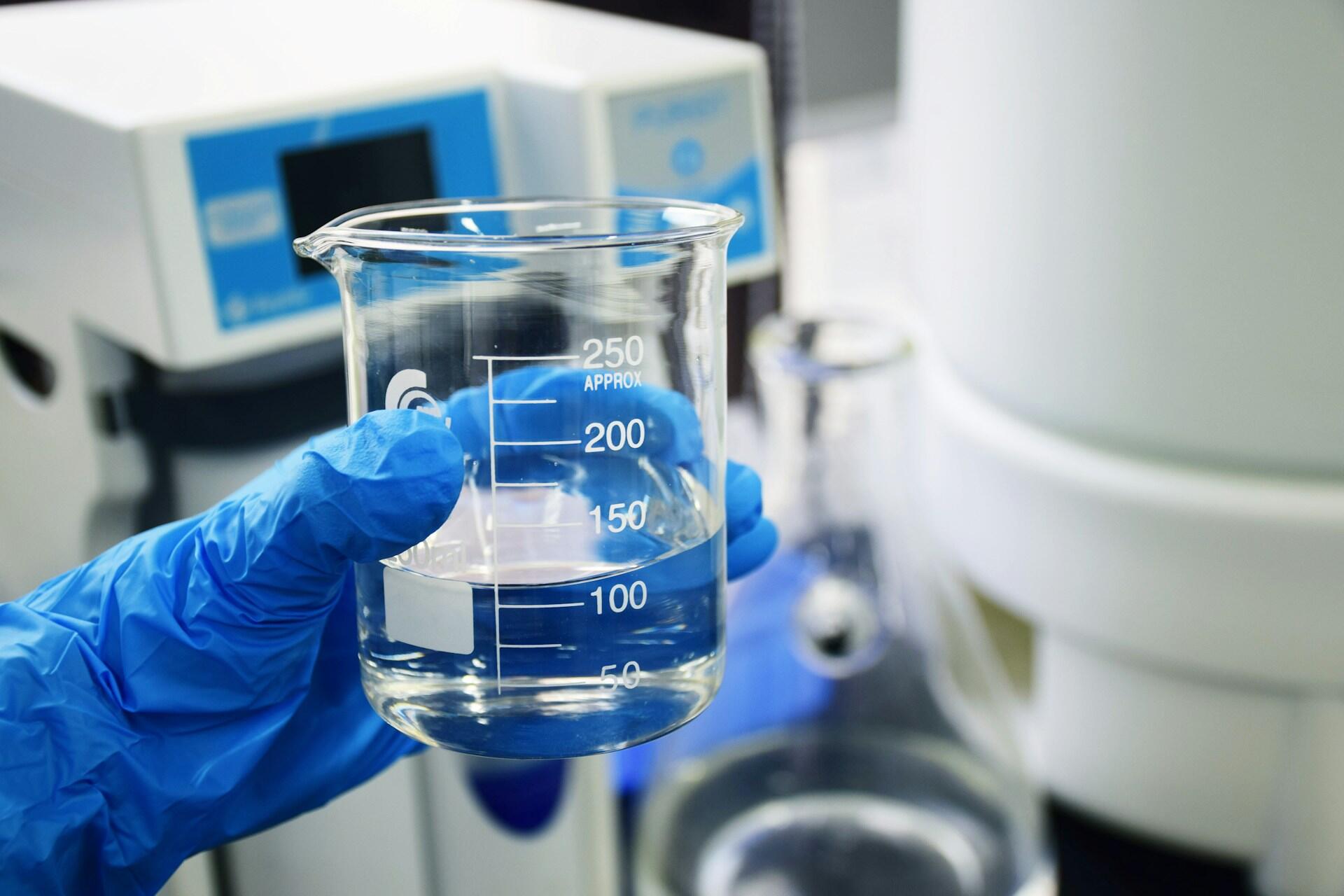 Inżynier chemik trzyma w dłoni w niebieskiej rękawiczce szklane naczynie laboratoryjne z płynem.