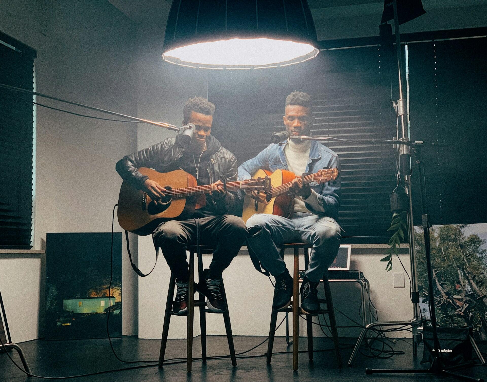 Dwóch mężczyzn siedzi na wysokich stołkach i gra na gitarach, oświetla ich duża lampa. Unsplash: Mika Chavala