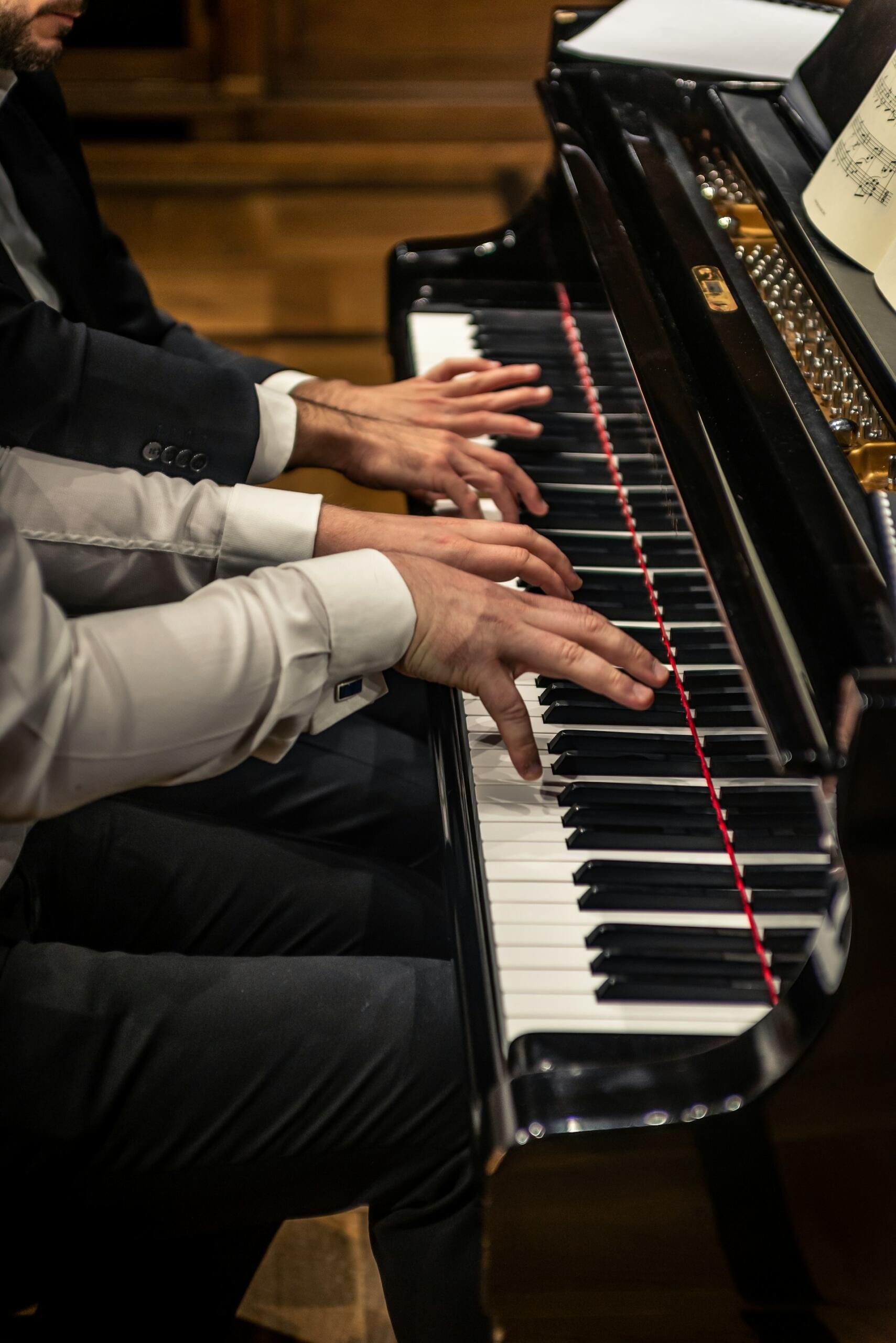 Dwóch elegancko ubranych muzyków wykonuje utwór na cztery ręce przy czarnym, lakierowanym fortepianie