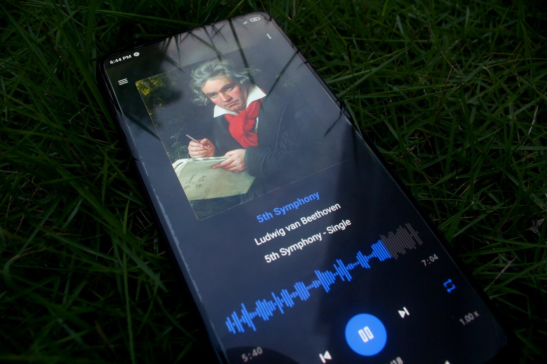 Smartfon leżący na zielonej trawie z włączoną aplikacją do odtwarzania muzyki, na wyświetlaczu odtwarzany utwór – „5th Symphony” (V Symfonia) z obrazem kompozytora Ludwiga van Beethovena.