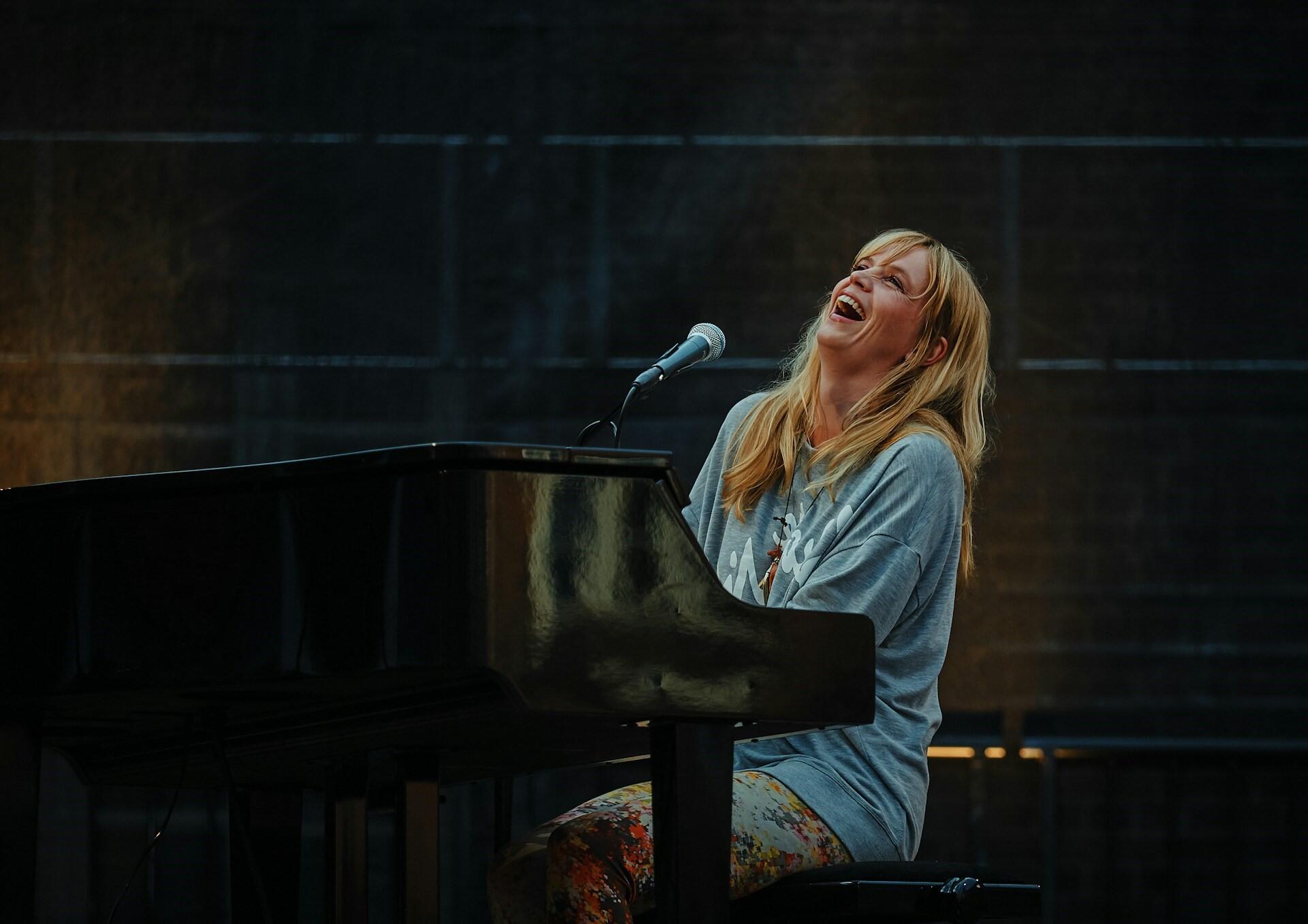 Uśmiechnięta wokalistka / pianistka, blondynka w luźnej bluzie i wzorzystych spodniach podczas występów na scenie. Unsplash: Vidar Nordli-Mathisen