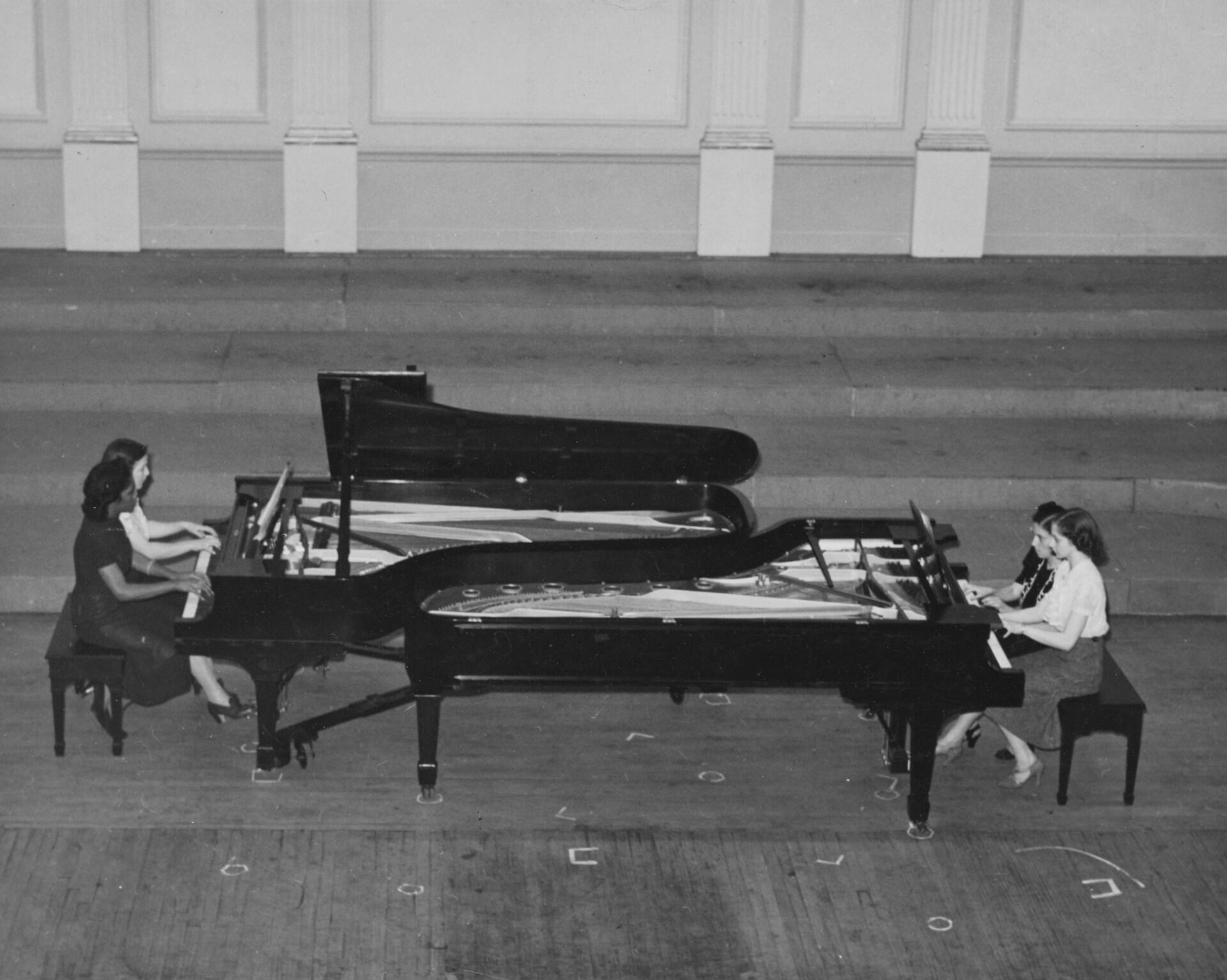 Cztery kobiety grające na dwóch fortepianach koncertowych usytuowanych naprzeciwko siebie (po dwie pianistki przy jednym instrumencie). Unsplash: The New York Public Library