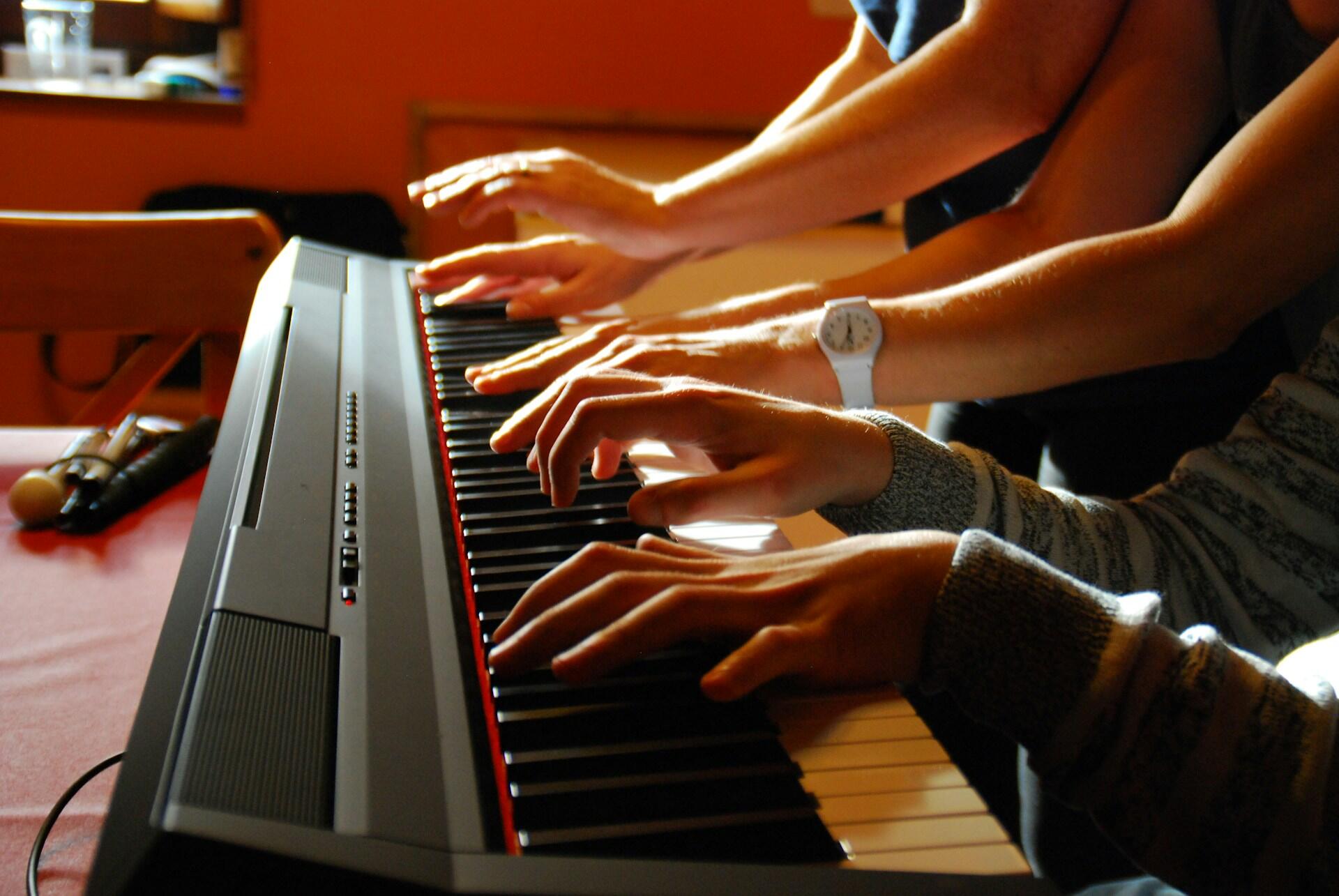 Dłonie kilku osób grających jednocześnie na jednym pianinie cyfrowym (keyboardzie). Unsplash: Miguel Saenz de Santa Maria