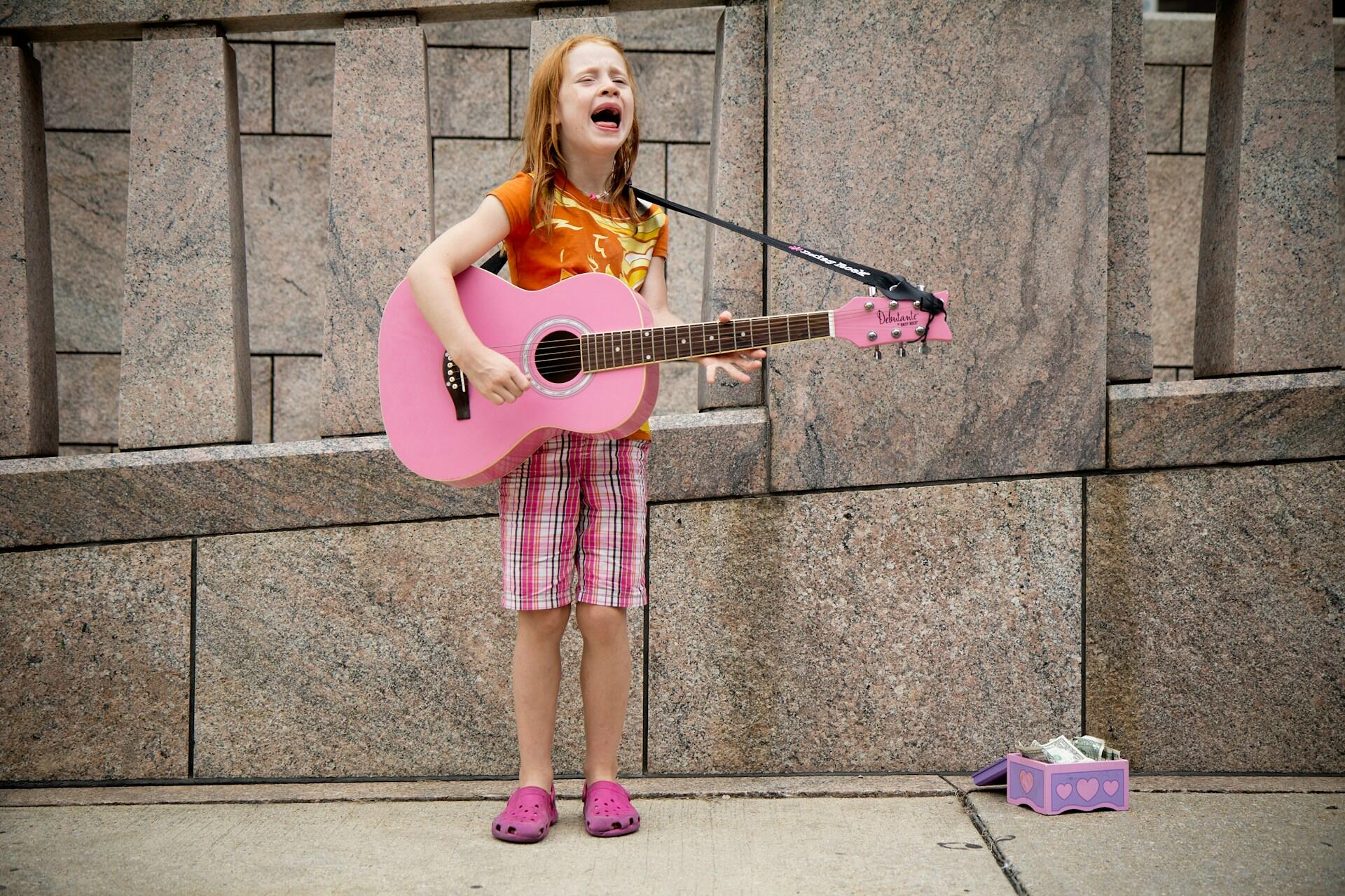 criança a cantar na rua com uma guitarra cor de rosa