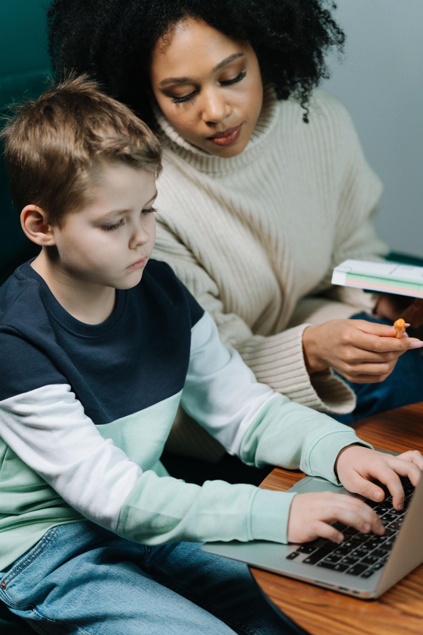 professora a apontar para livro enquanto aluno escreve em computador