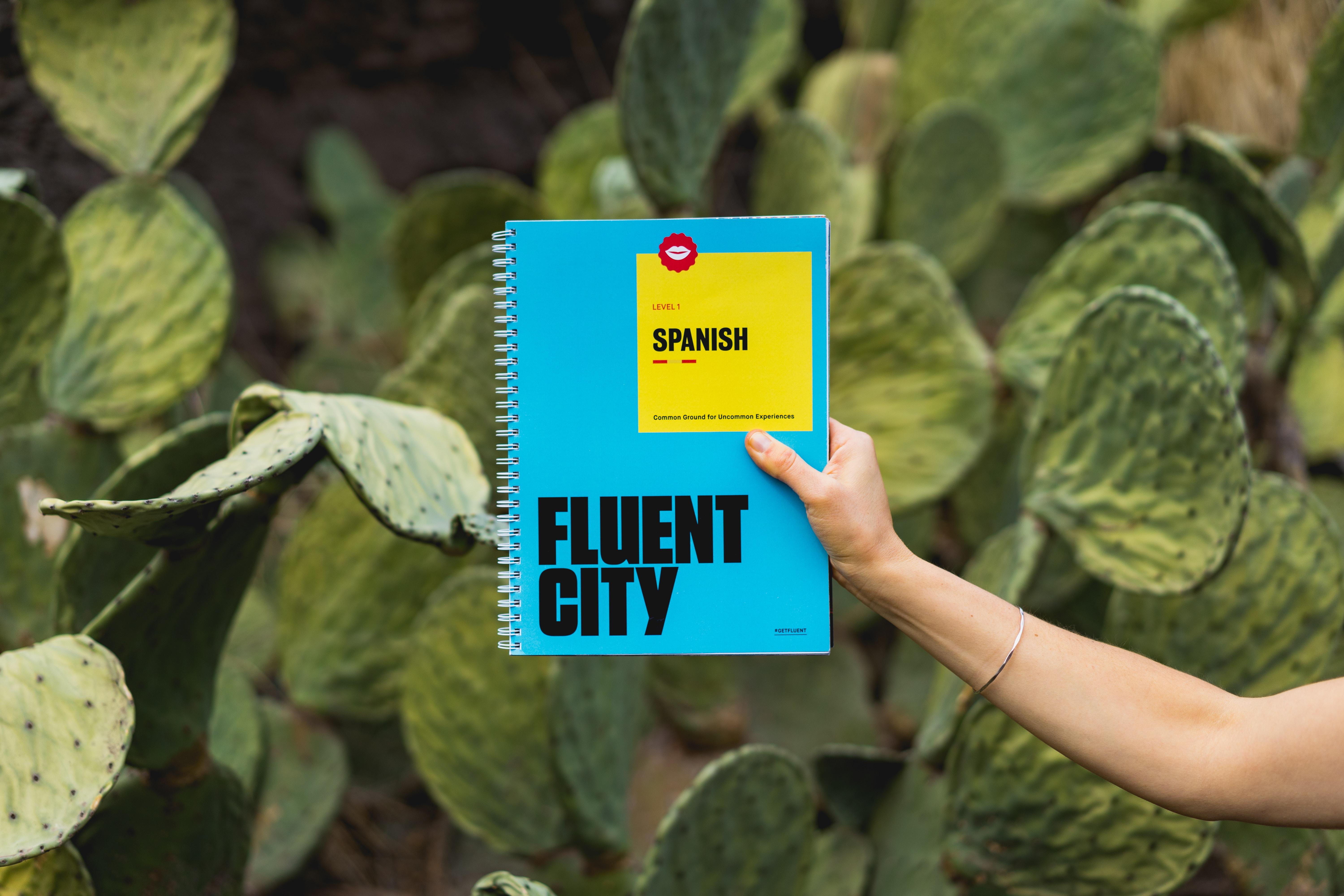Uma pessoa a segurar um livro de espanhol frente a um fundo de vegetação.