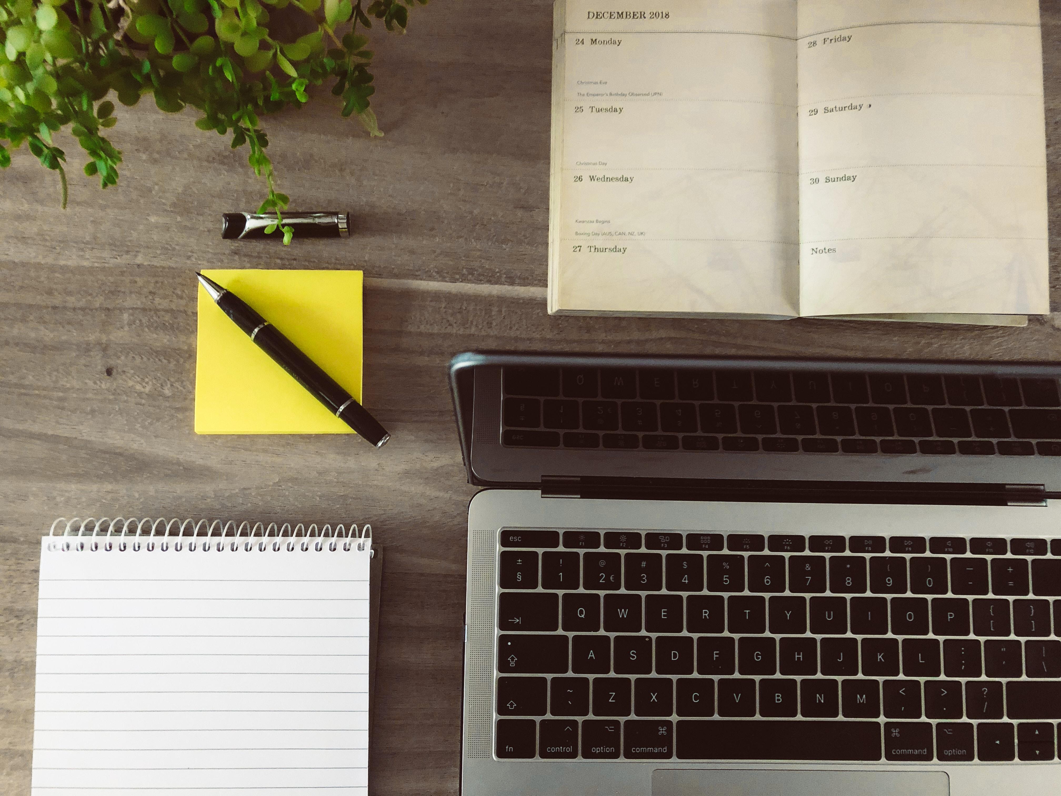 Um computador, uma caneta, post-its e uma agenda de um programador cultural em cima de uma secretária.