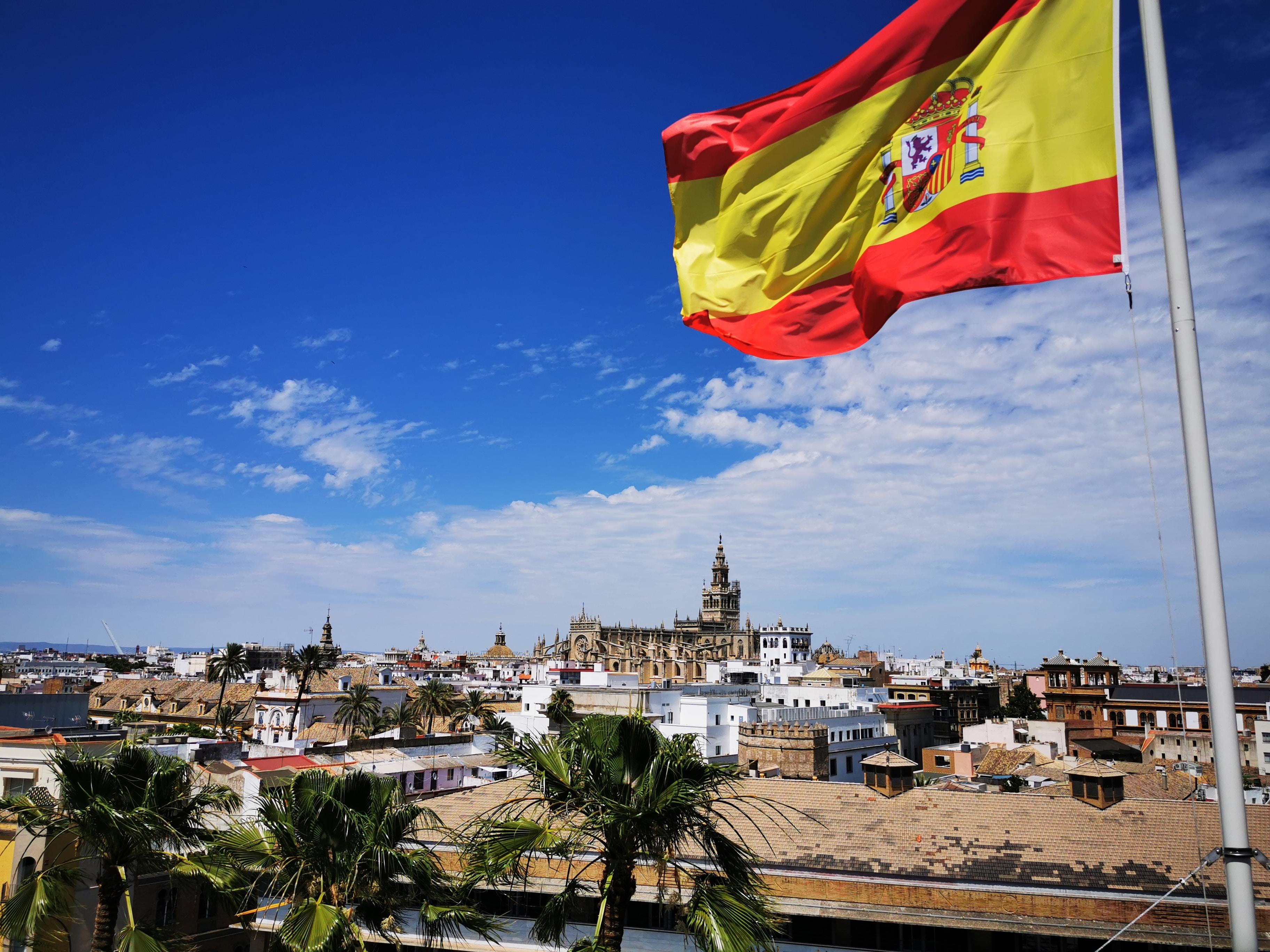 Uma bandeira de Espanha, ao vento, com a paisagem de uma cidade.