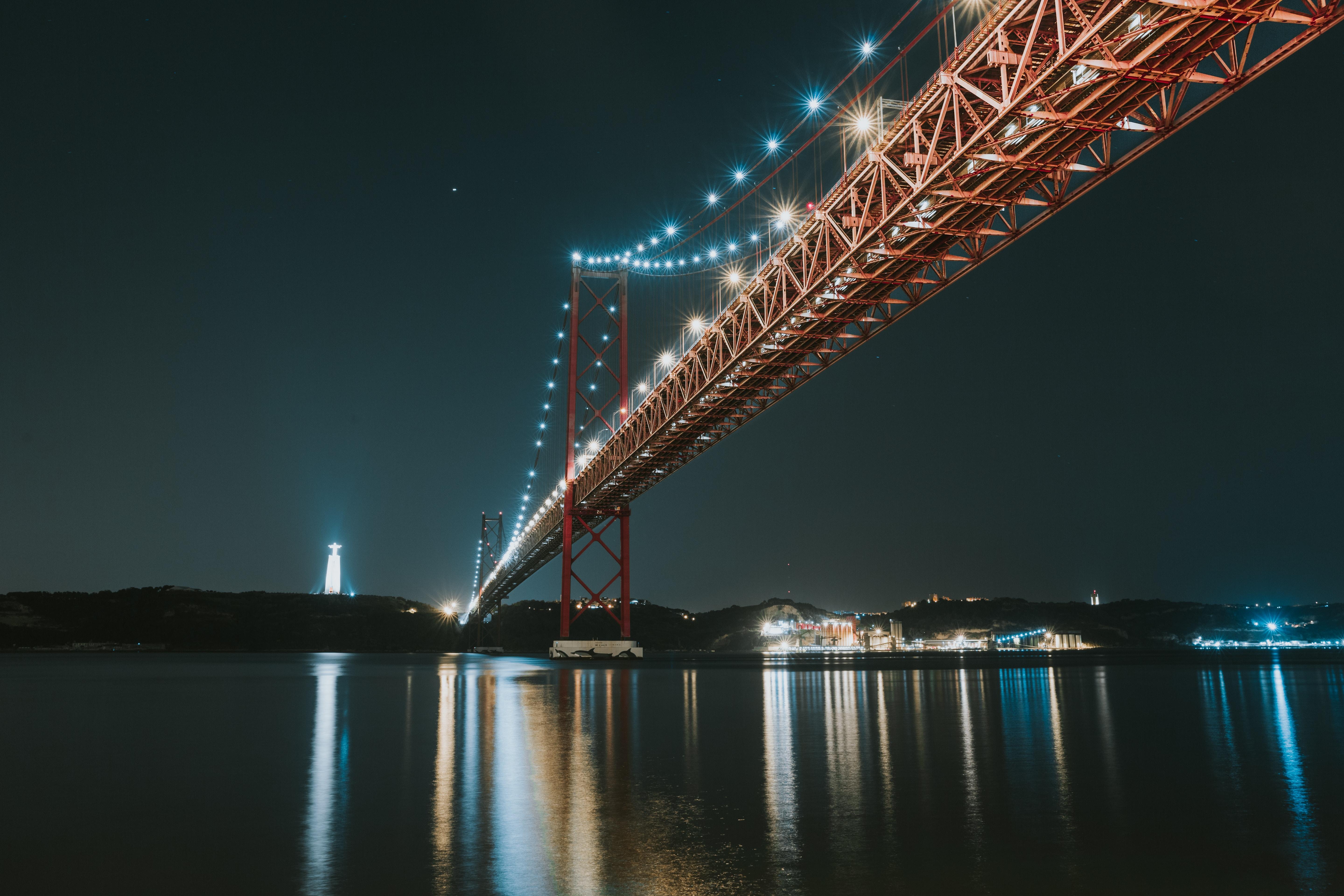 Uma fotografia tirada da margem norte do rio Tejo, com a ponte 25 de Abril e o Cristo Rei iluminados, à noite.