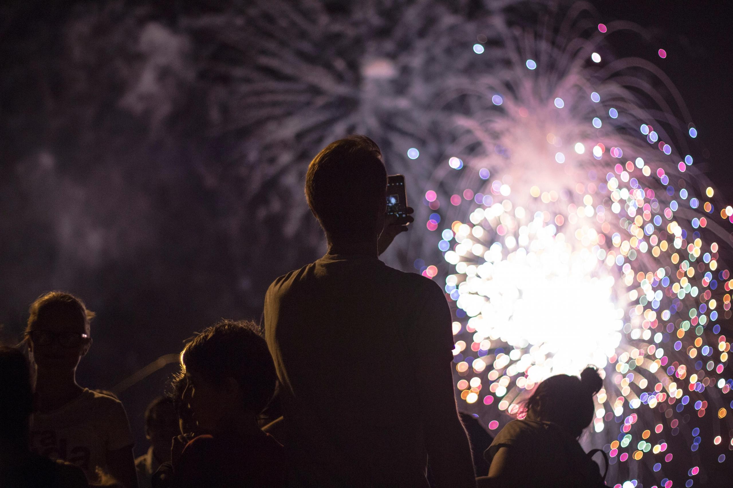 Várias pessoas a assistir e um rapaz a filmar um espetáculo de fogo de artifício