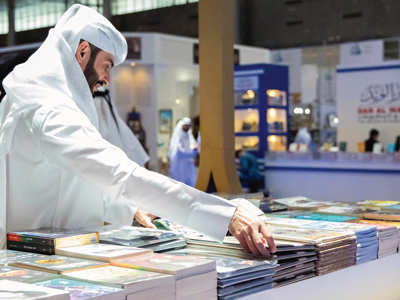رجل يرتدي الزي القطري التقليدي يتصفح مجموعة من الكتب المعروضة في جناح داخل معرض الكتاب.